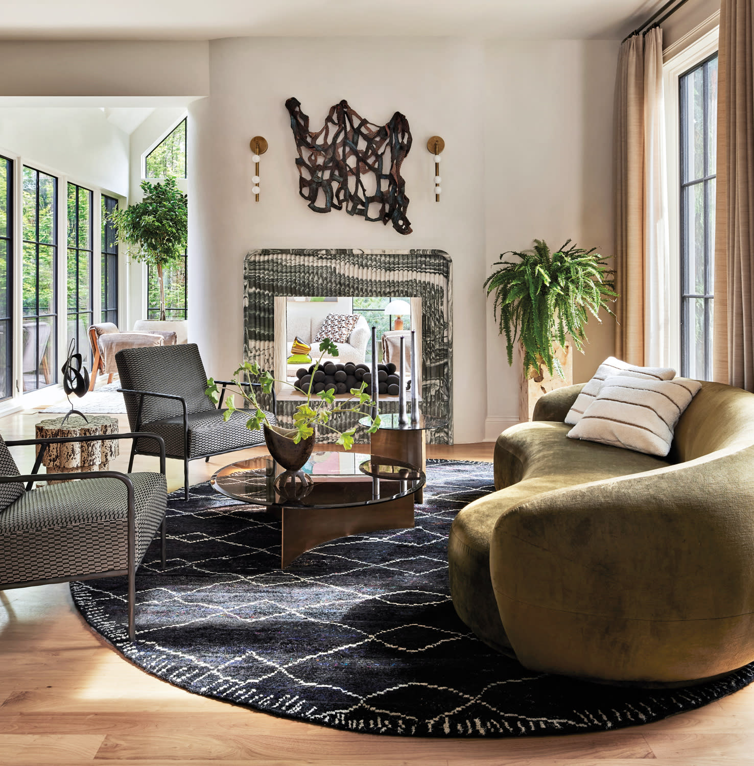 Modern living room with a green velvet chair, black geometric rug, and glass coffee table. Large windows and plants add a bright, airy feel.