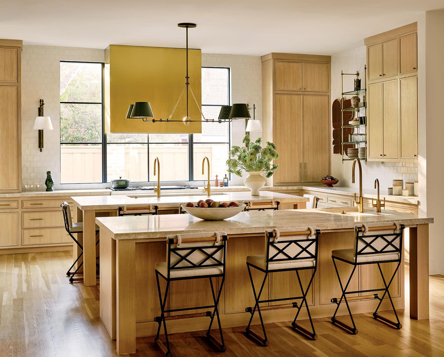 Spacious kitchen with light wood cabinetry and flooring, featuring a large island with black metal chairs. A yellow range hood adds a pop of color.