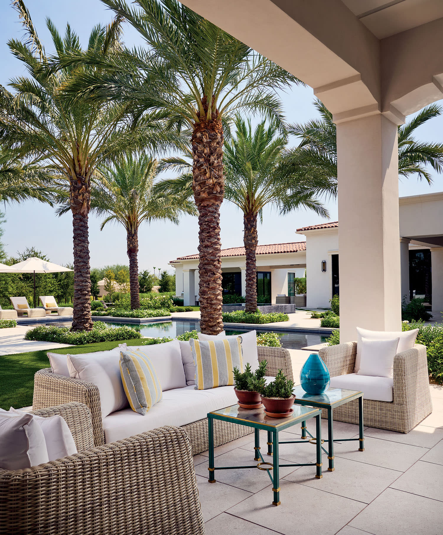 Outdoor patio with wicker furniture and striped pillows overlooks a lush garden with palm trees. Calm ambiance, modern villa in the background.