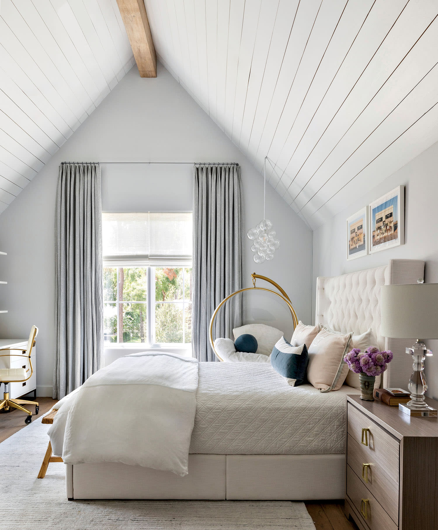 Bright, modern bedroom with high, white-paneled ceiling and wooden beam. Features elegant bed with pastel pillows, gold-accented chair, and side table with flowers.
