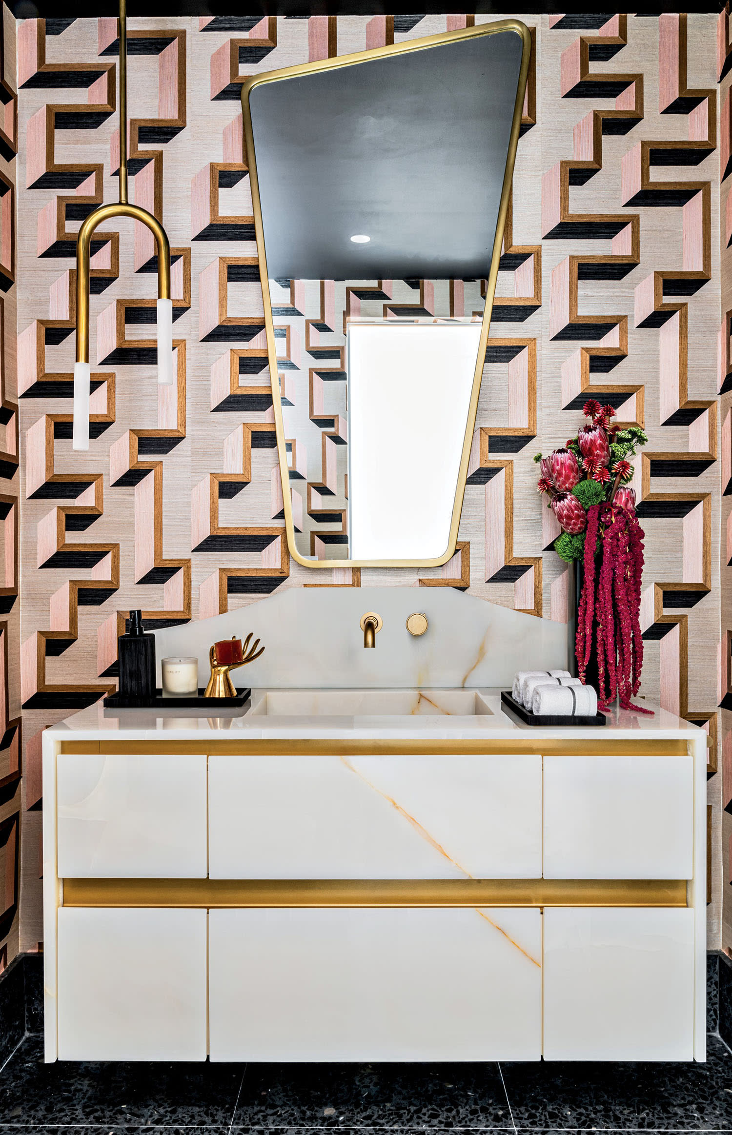 Trendy bathroom with geometric patterned wallpaper, angular mirror, gold fixtures, and a white vanity. Elegant flowers add a touch of color.