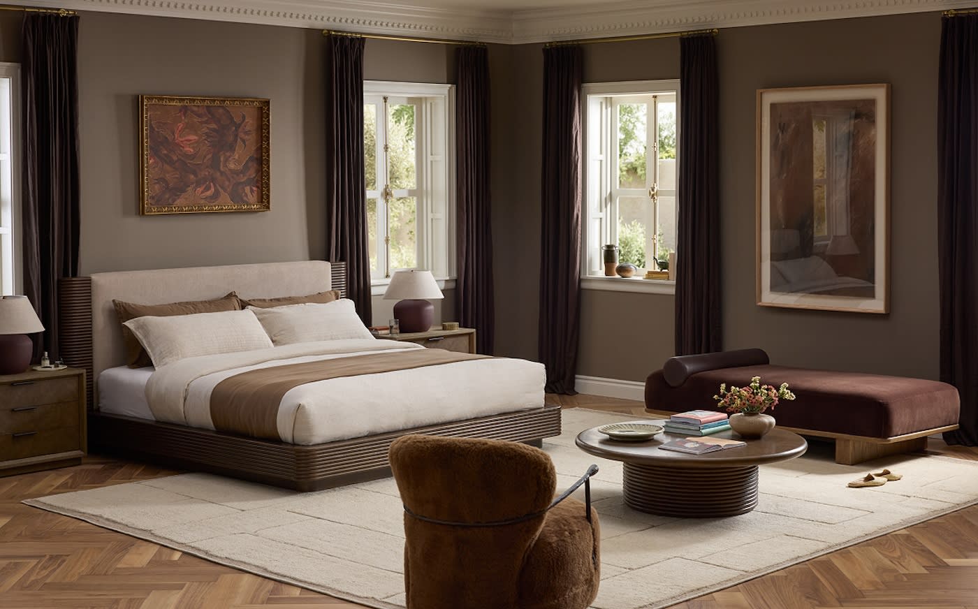 Sophisticated bedroom with a brown and cream color scheme. Features a large bed, chaise lounge, cozy chair, round coffee table, artwork, and tall windows.