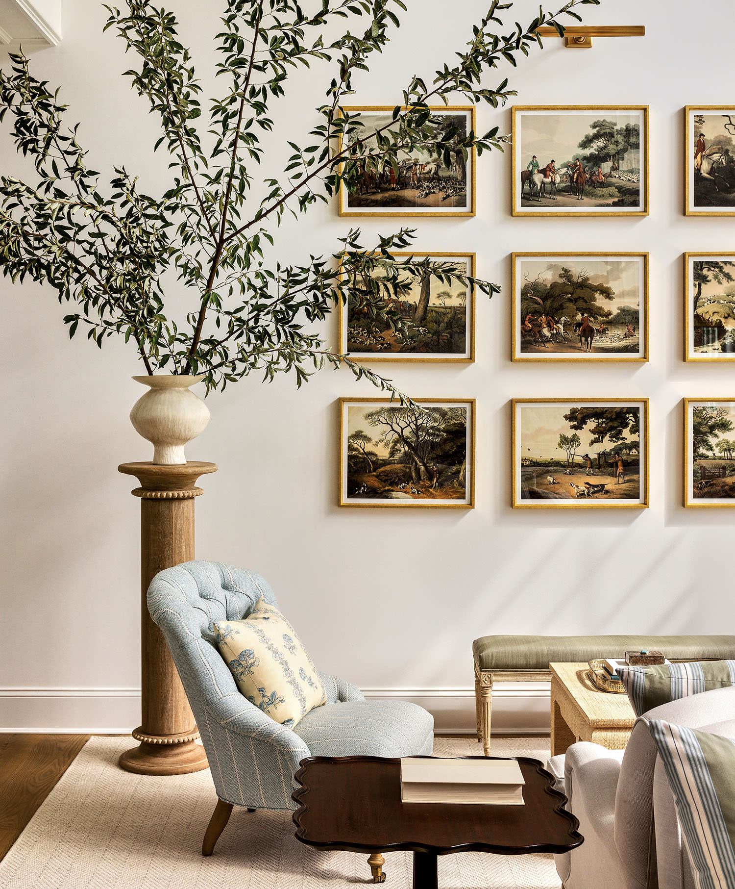 Elegant living room with a tall potted plant, light blue tufted chair, small wooden table, and framed artwork arranged in a grid on the wall.