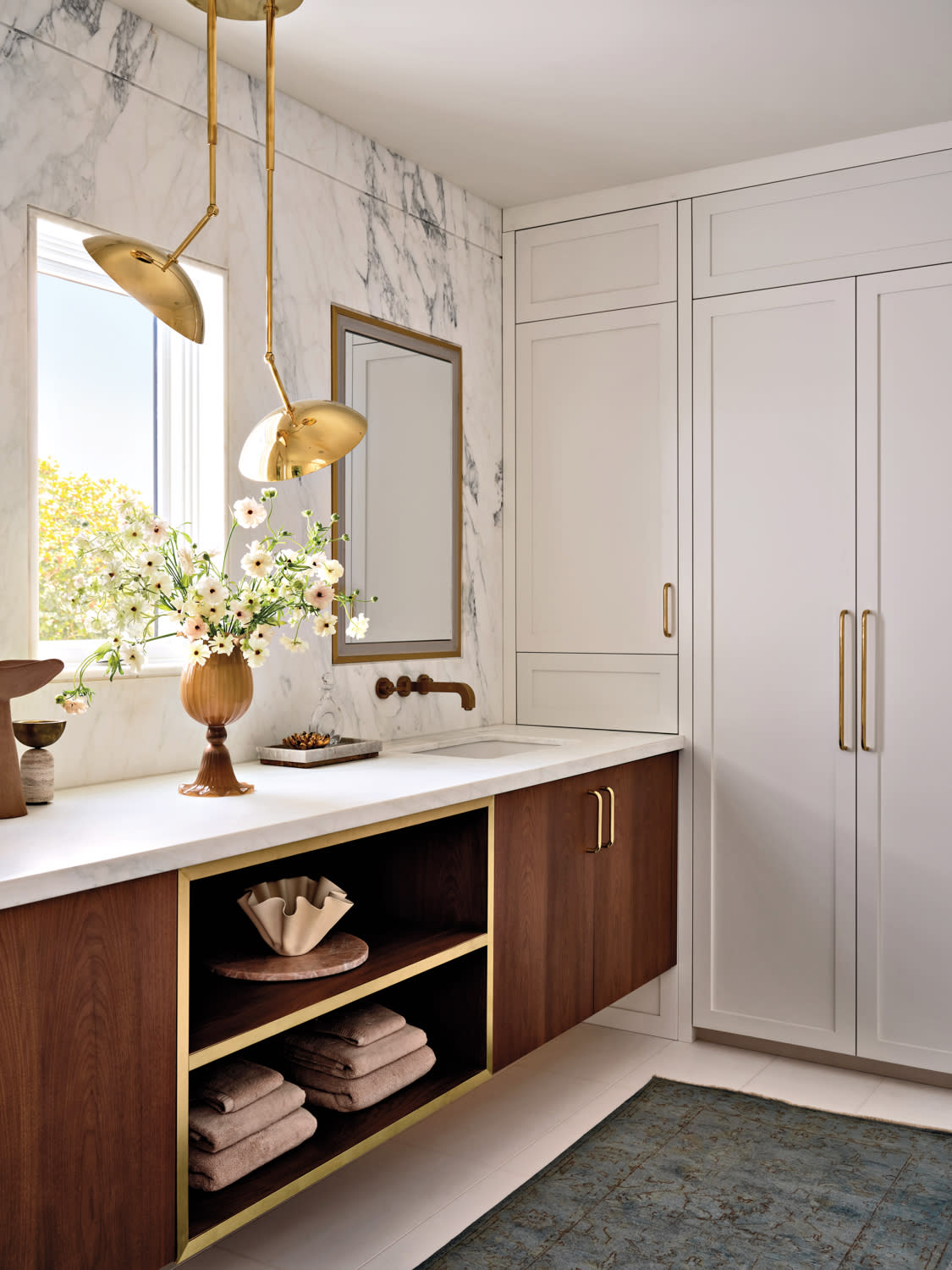 Stylish bathroom with marble walls, wooden vanity, bronze fixtures, and floral arrangement. Soft light from a window adds warmth and elegance.