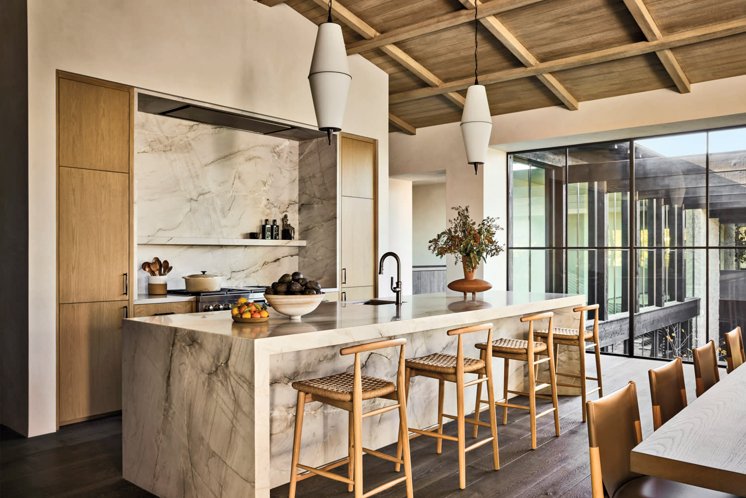 Modern kitchen with marble island, wooden stools, and pendant lights. Large windows provide natural light. The setting is elegant and inviting.