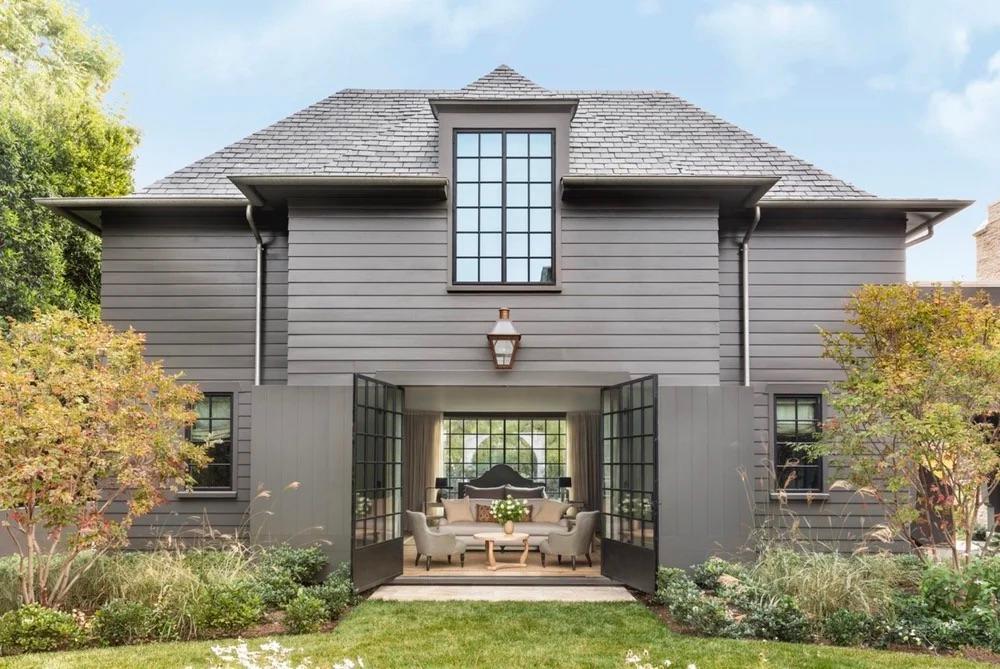 Gray two-story house with large windows and open French doors, revealing a cozy living area. Surrounded by lush greenery under a clear blue sky.