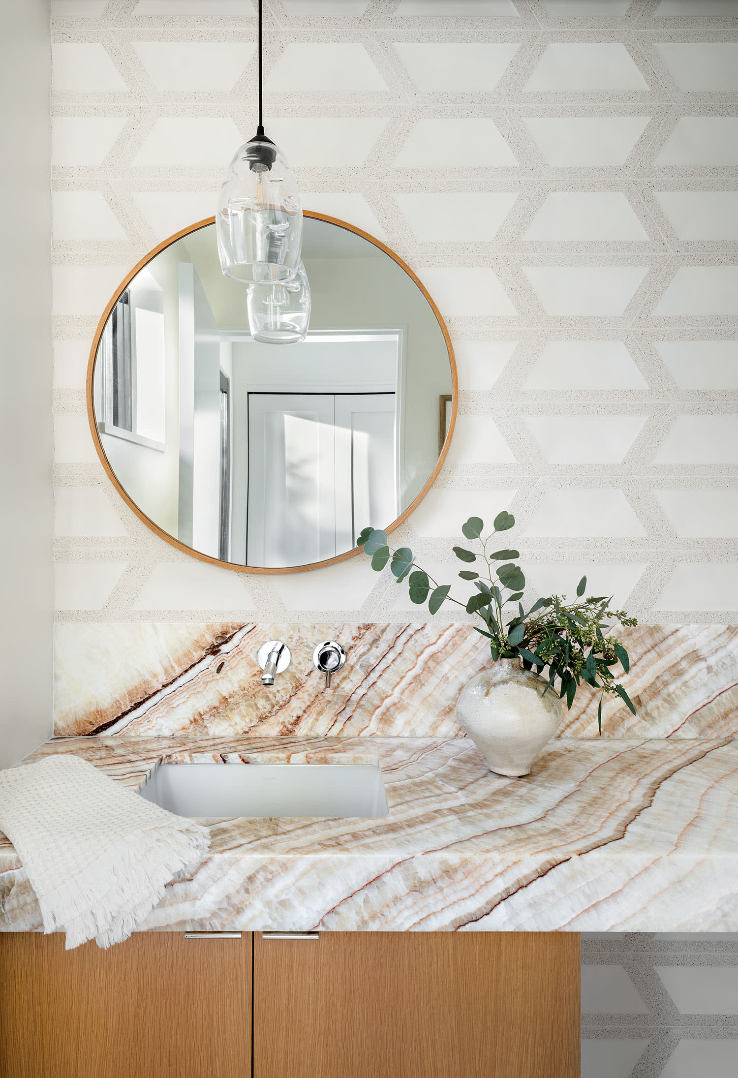 Modern bathroom with hexagonal wallpaper, a round mirror reflecting a doorway, and a marble countertop. A vase with greenery adds a fresh touch.