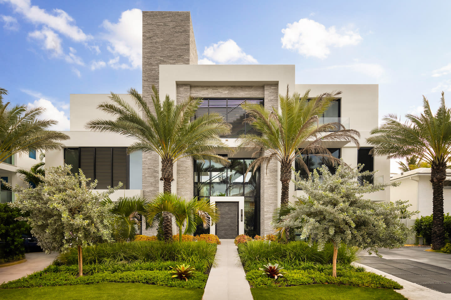 Contemporary coastal mansion composed of crisp white volumes and stacked gray-stone towers, its double-height glass entry framed by slim palms and charcoal louvered windows. A straight concrete walkway bisects lush, low-slung tropical landscaping—silverleaf trees, bromeliads, and manicured lawn—guiding the eye to a deep slate front door beneath a bright blue, cotton-cloud sky.