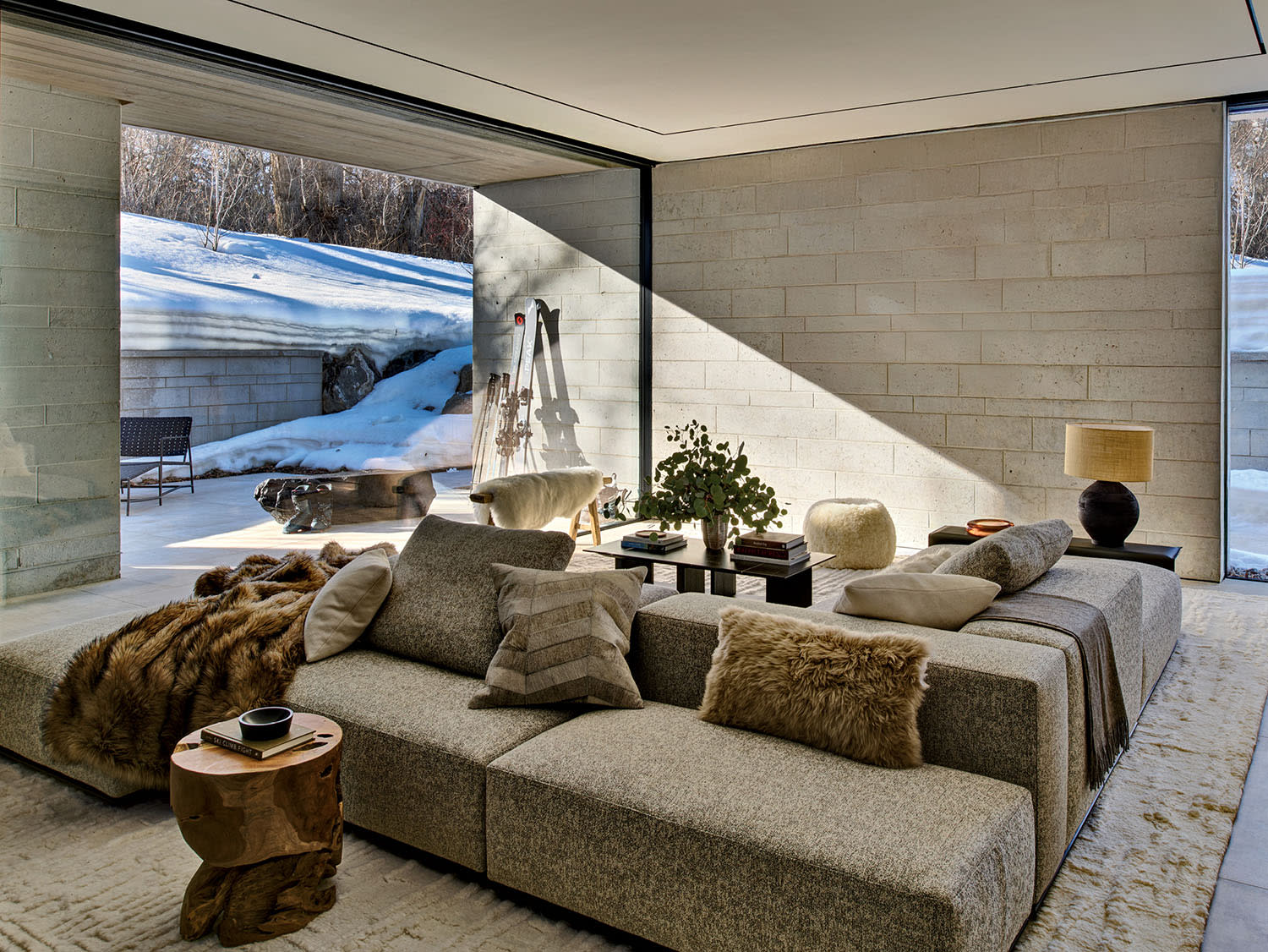 Modern living room with floor-to-ceiling windows, natural light on gray sofas, fur accents, and wooden decor.