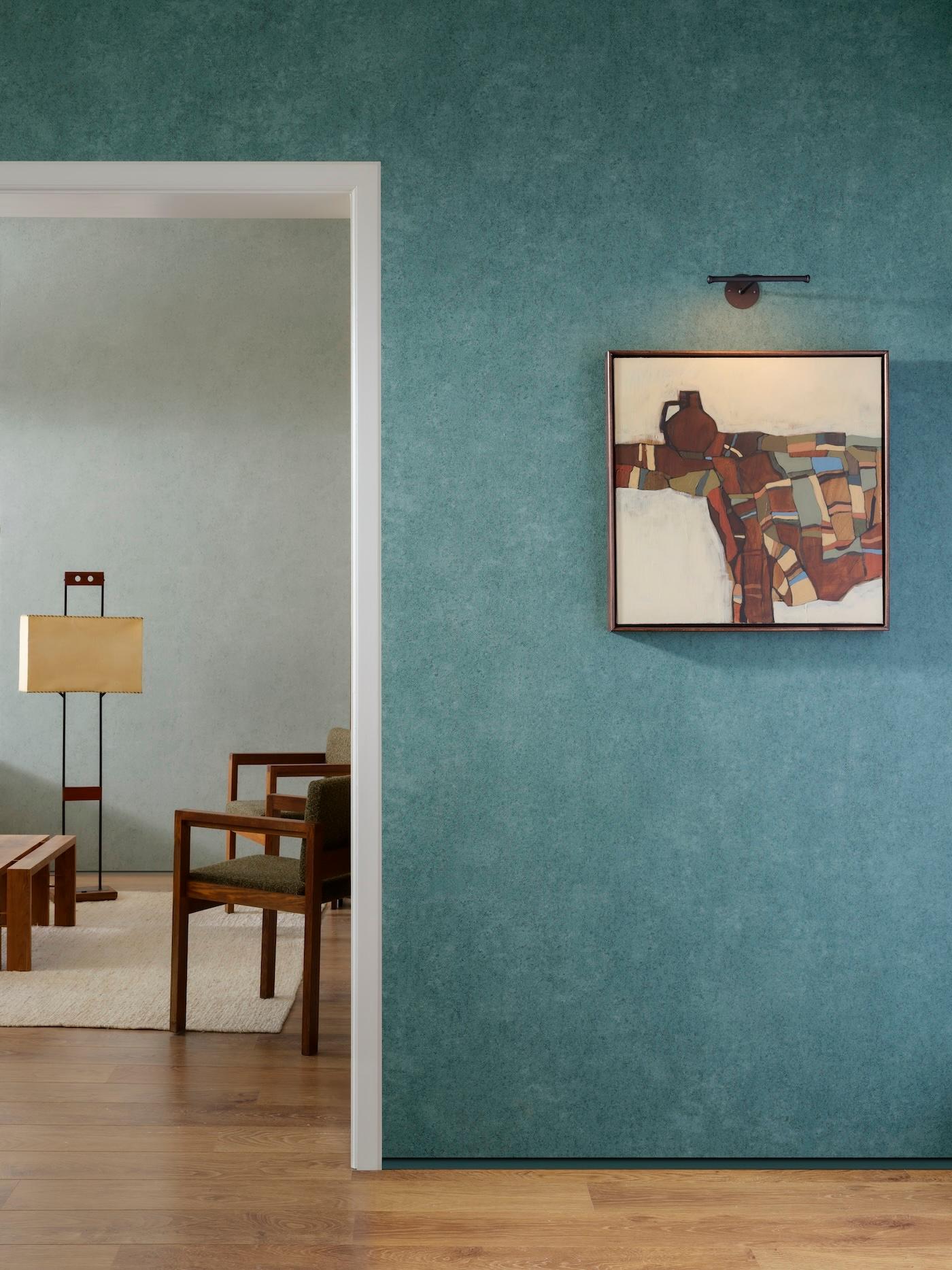 Contemporary room with blue-green walls, featuring abstract art. Through a doorway, a cozy seating area is visible, furnished with a lamp and chairs, creating a serene atmosphere.