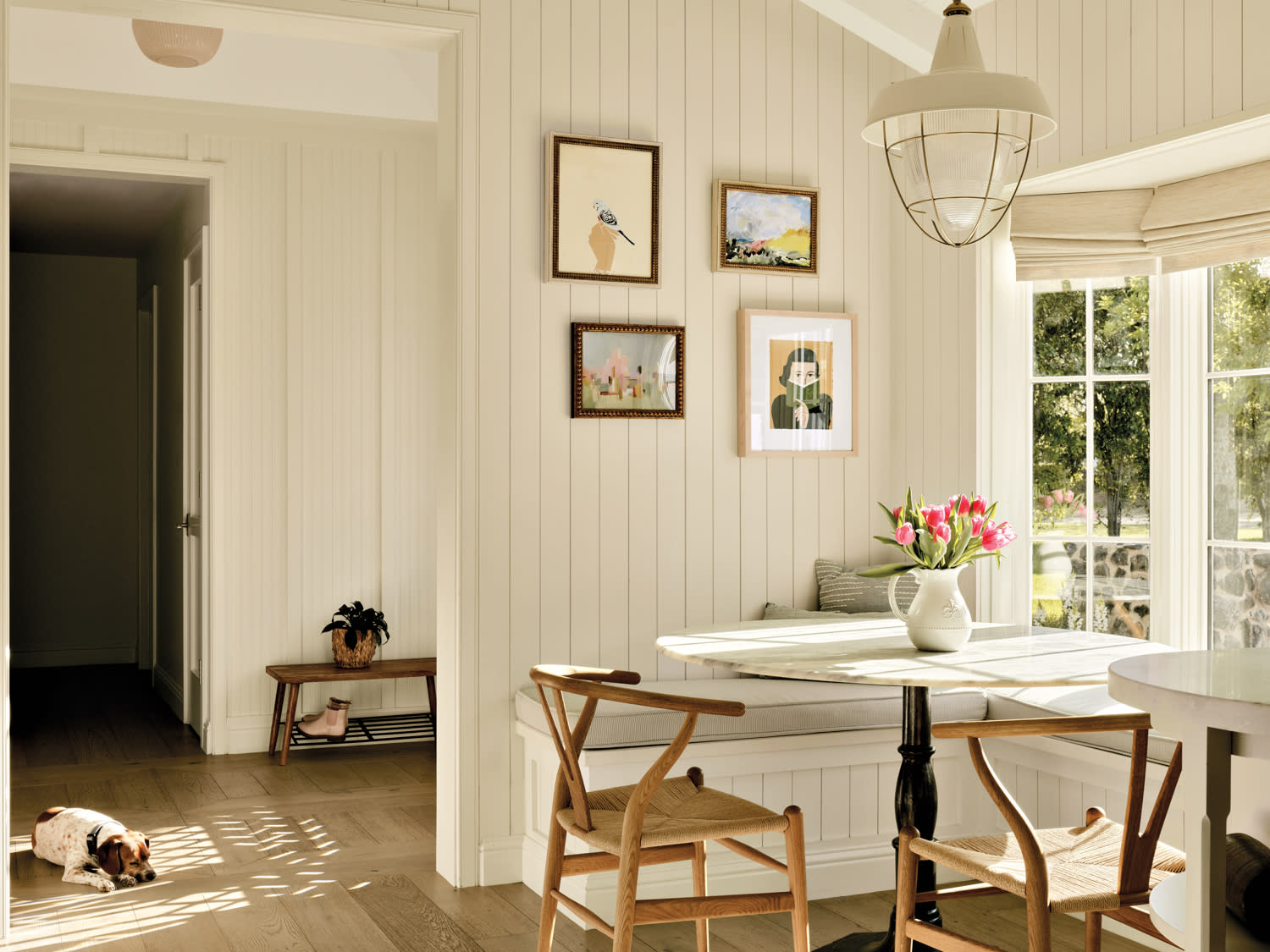 Four small paintings hang above a corner banquette and round table next to a kitchen window