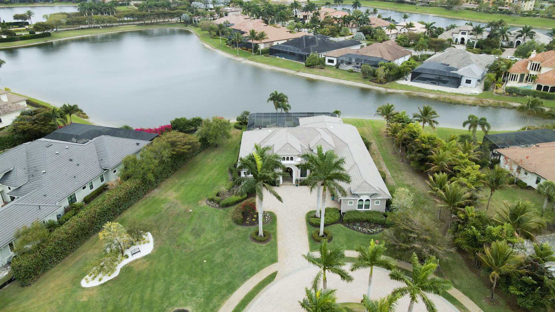 Aerial view of a large, luxurious house with a well-maintained lawn and palm trees. It sits by a tranquil lake, surrounded by other upscale homes.