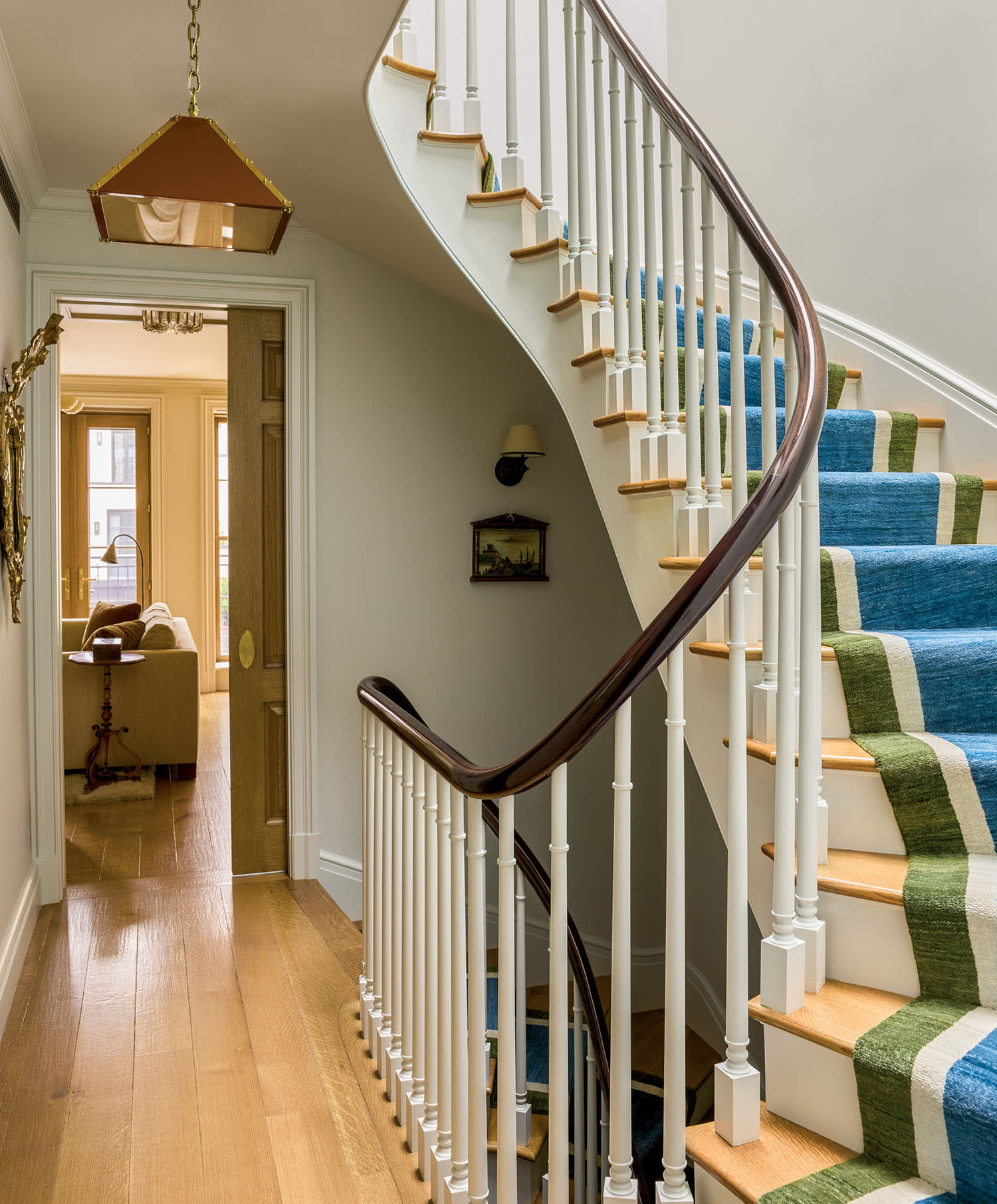 Curved staircase with blue-and-green striped carpet and wooden railing. Warm-lit hallway with hardwood floor leads to a cozy living room with sunlight.
