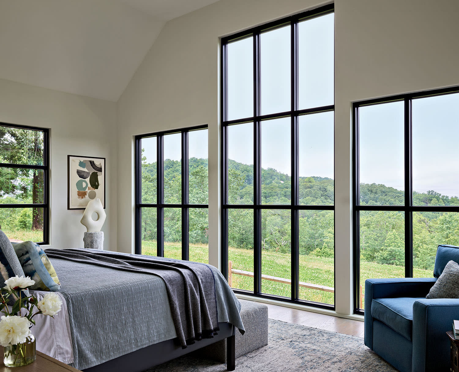 A bedroom made up with a bed and armchair, with artwork in a corner and large windows.