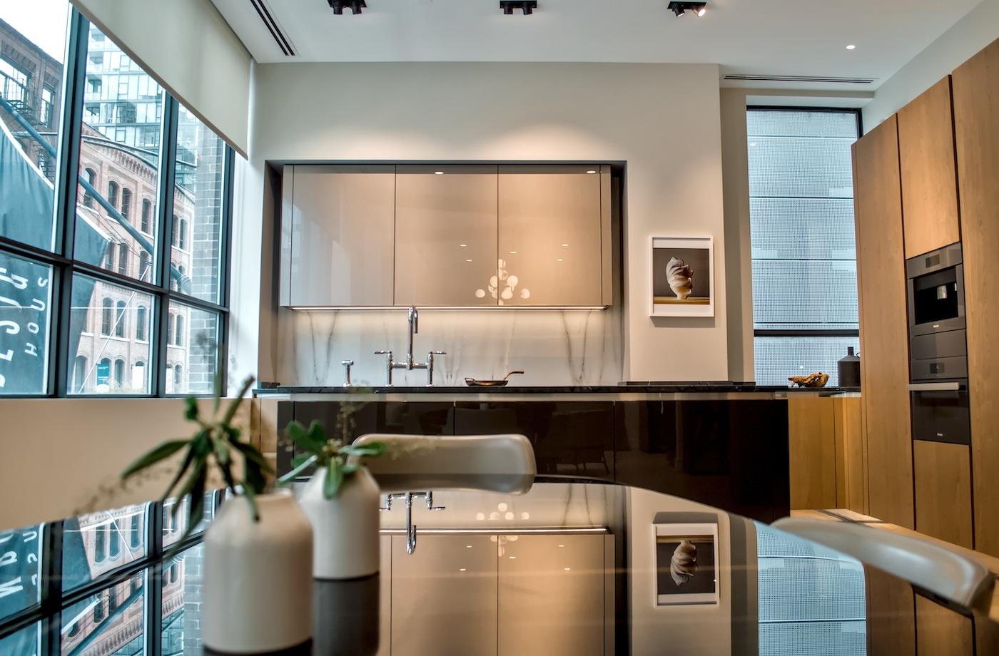 Modern kitchen with sleek cabinetry, built-in appliances, and large windows showing a cityscape. Reflections on a glossy table add elegance.