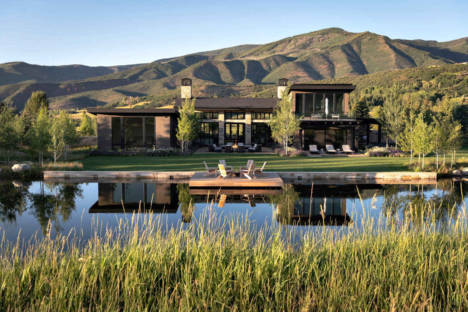 A modern home beside a tranquil pond, with majestic mountains rising in the background under a clear blue sky.
