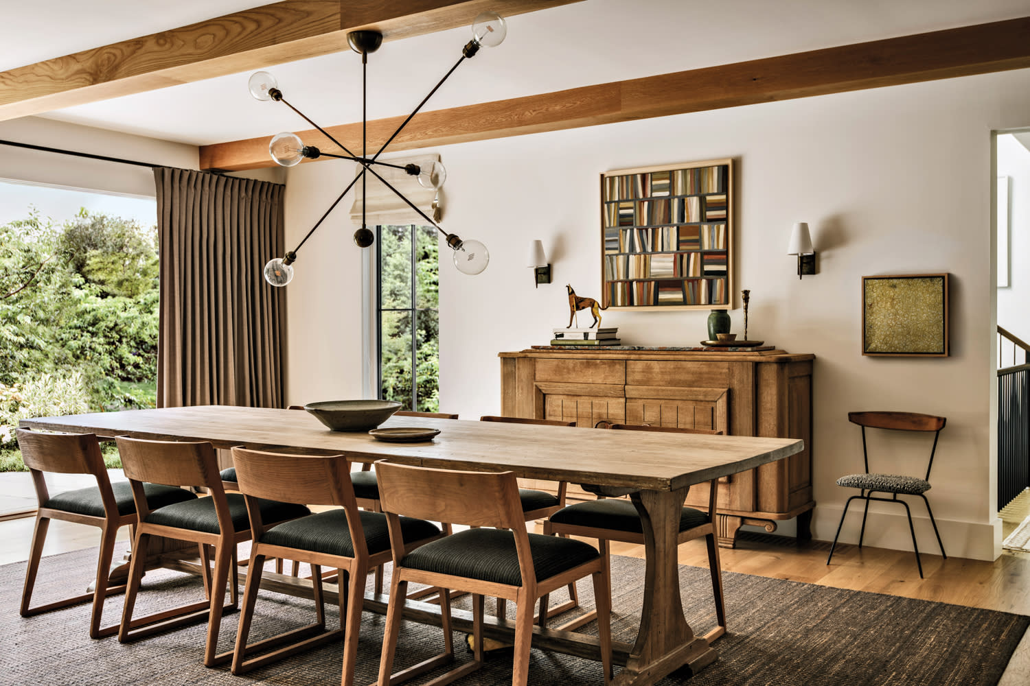 Stylish dining room with a long wooden table, six chairs, and a modern chandelier. Large window with curtains and greenery outside add warmth.