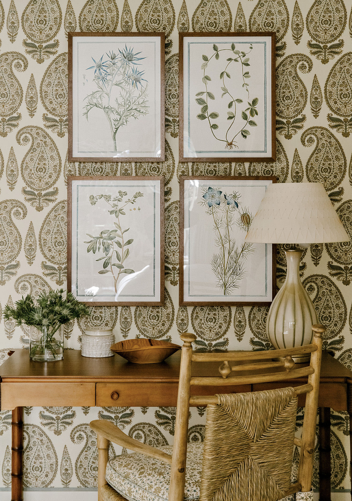 Wallpaper with a paisley print on a wall with four framed photos of plants set over a desk and chair.