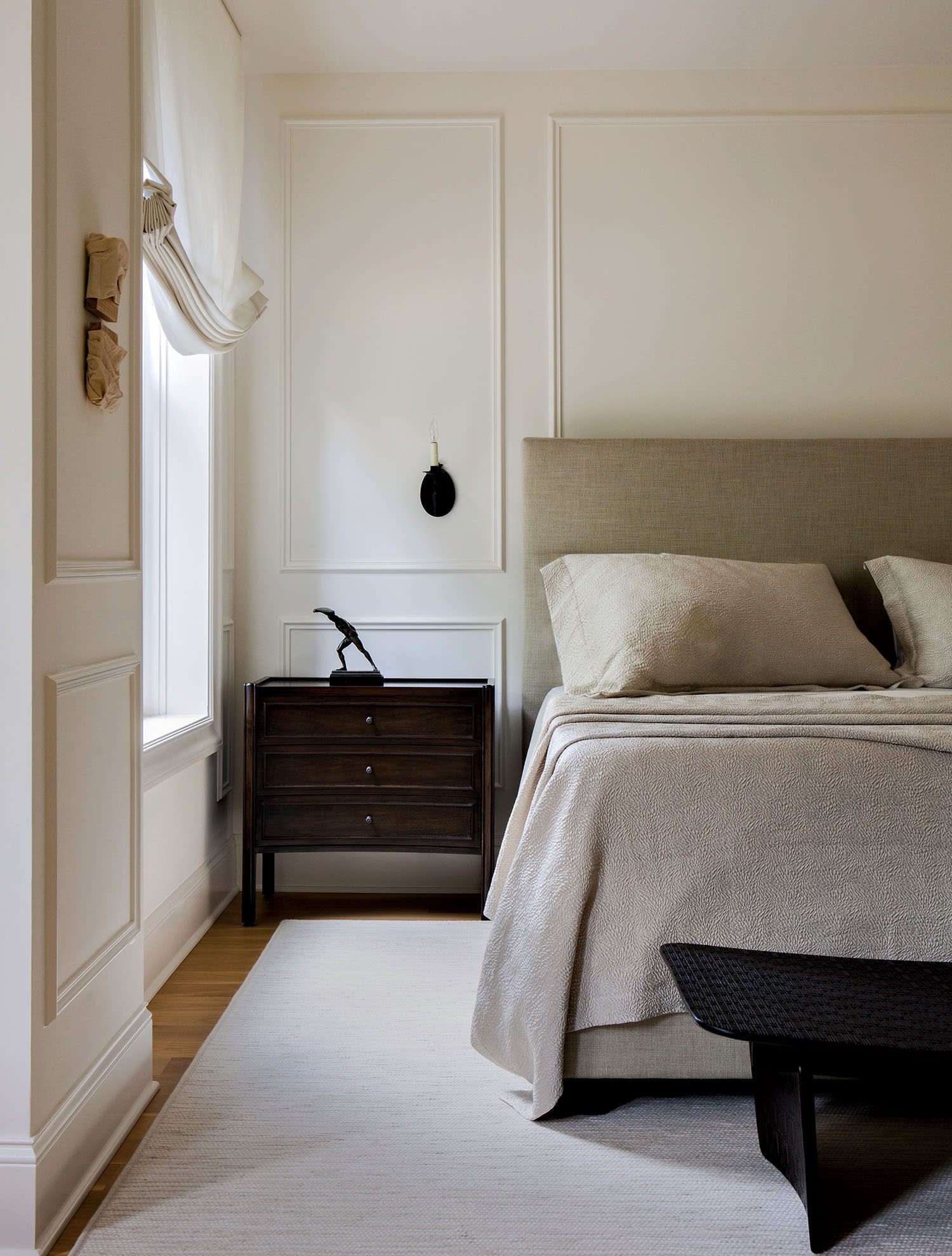 A bed with an upholstered headboard sits next to a dark wood nightstand topped with a small figurine.