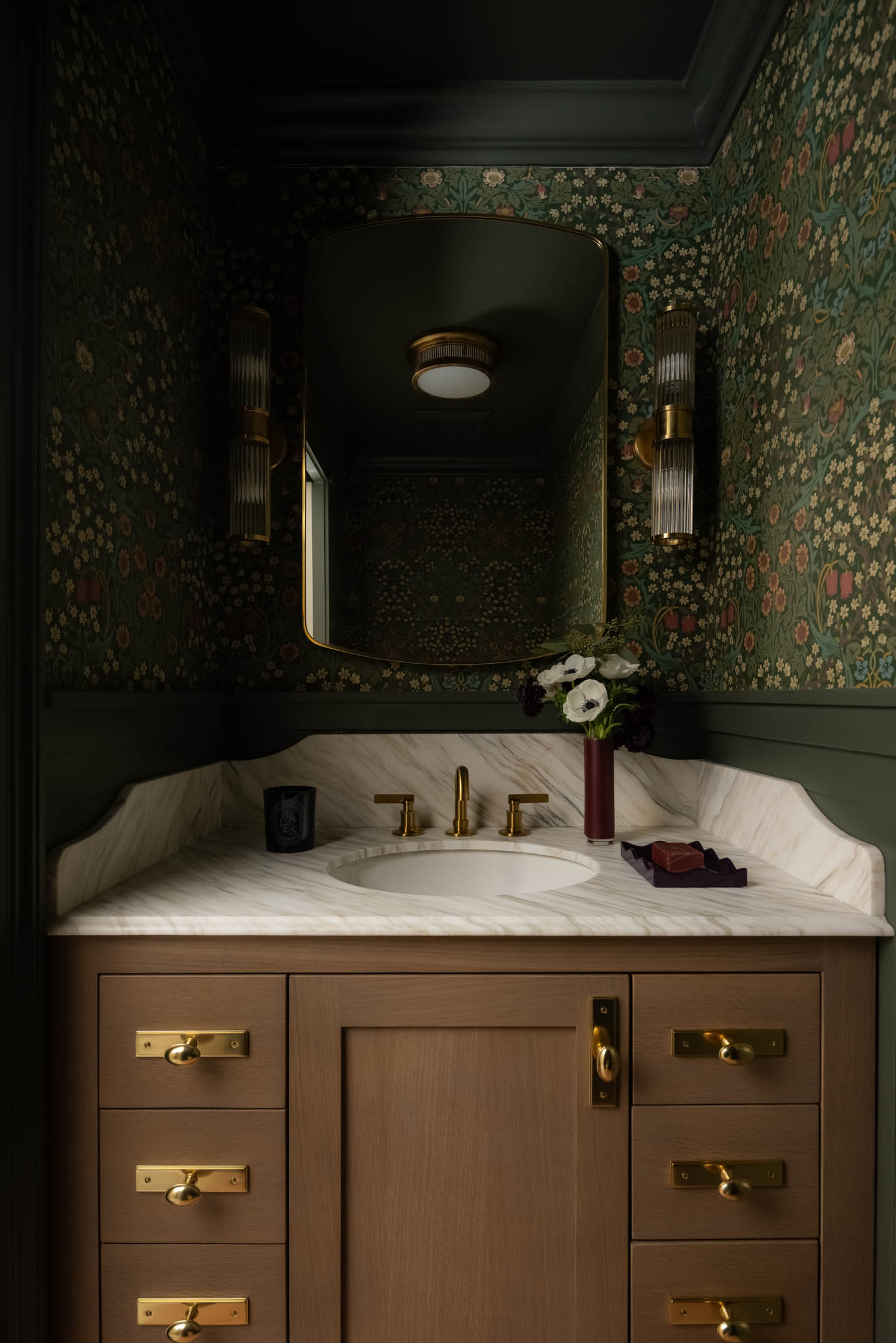 Dark, floral-patterned bathroom with wood vanity and brass fixtures. A round mirror and sconces hang above a marble sink, exuding a vintage, elegant vibe.