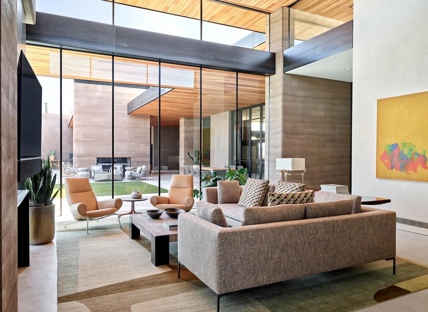 Modern living room with beige sofa, geometric cushions, and two tan armchairs facing a coffee table. Glass wall reveals patio and garden. A vibrant abstract painting adorns the white wall, adding a splash of color. The room feels spacious and serene.