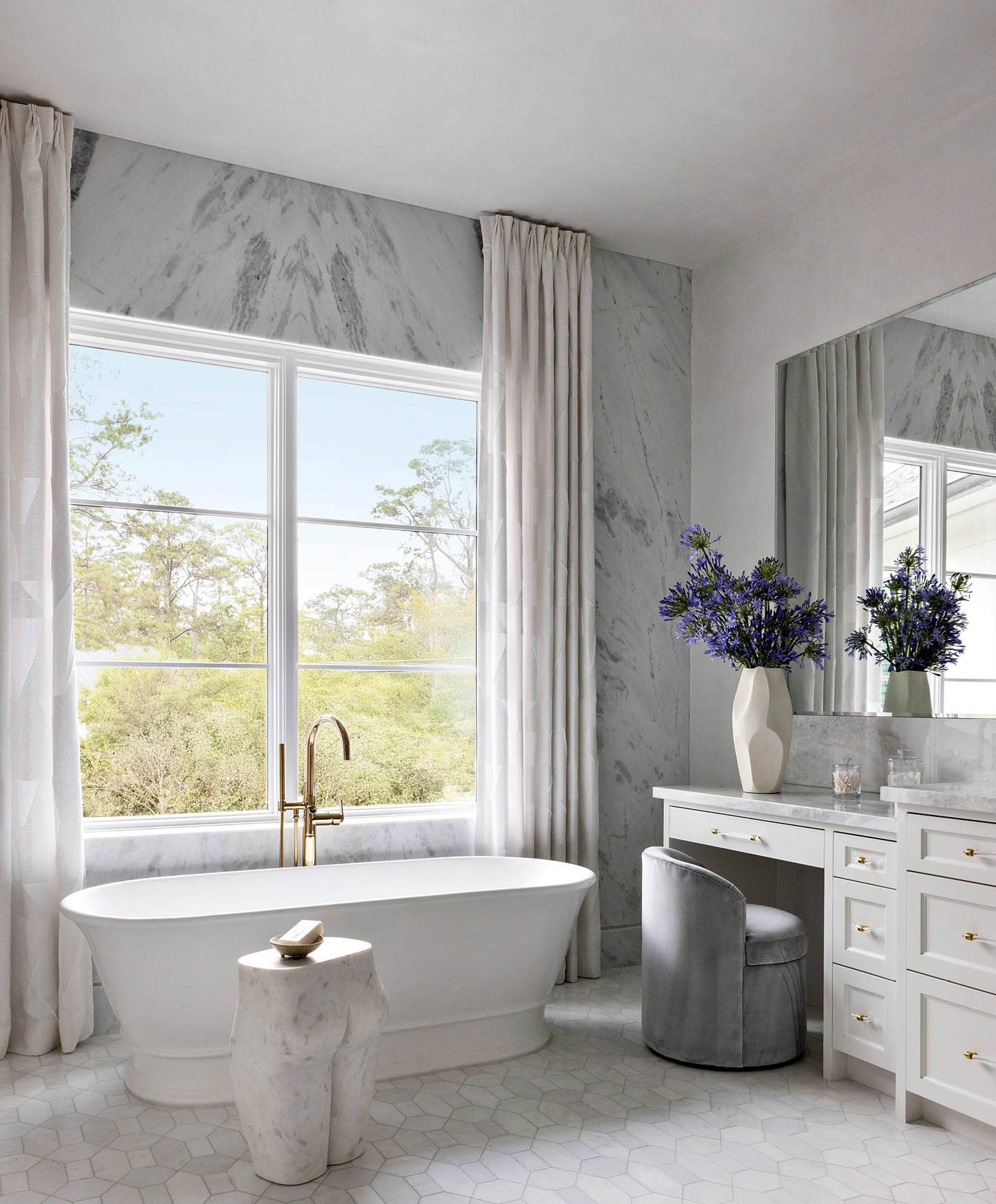 A freestanding bathtub sits under a window near a side table, a white vanity and gray chair.