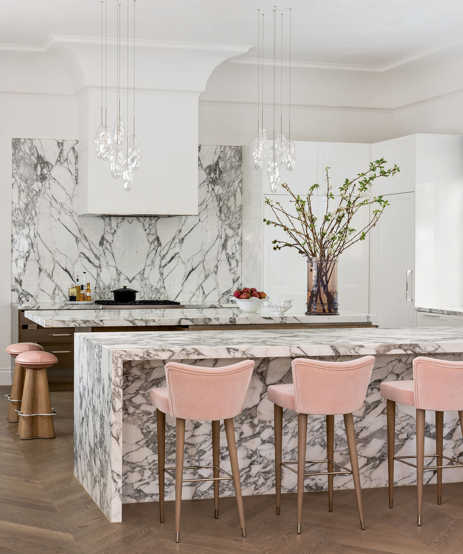 Luxurious kitchen with marble countertops, a matching backsplash, and modern pendant lights. Pink bar stools add a soft touch. Vase with branches decorates the counter.