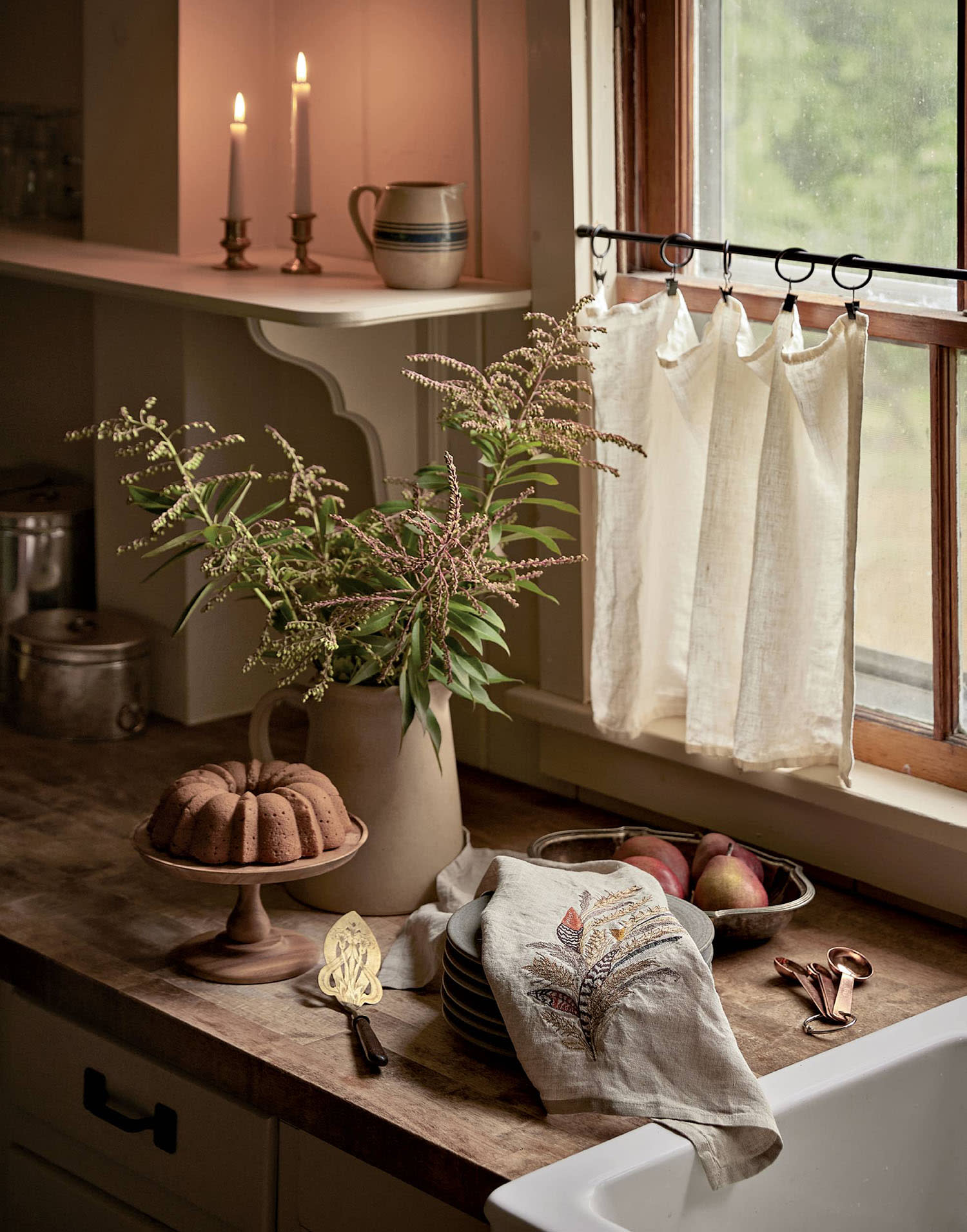 A kitchen counter with a bundt cake on a cake stand, greenery in a vase and a bowl of fruit.