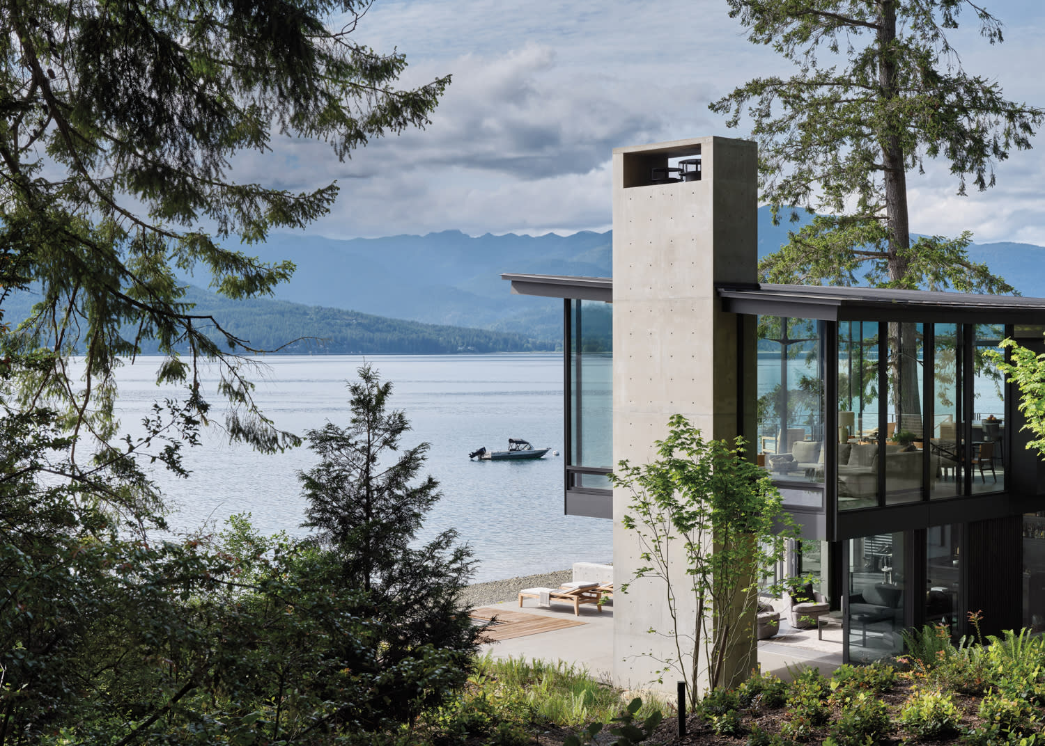 Modern lakeside house with large glass windows and concrete structure, surrounded by trees. A boat floats on the calm water, mountains in the background.