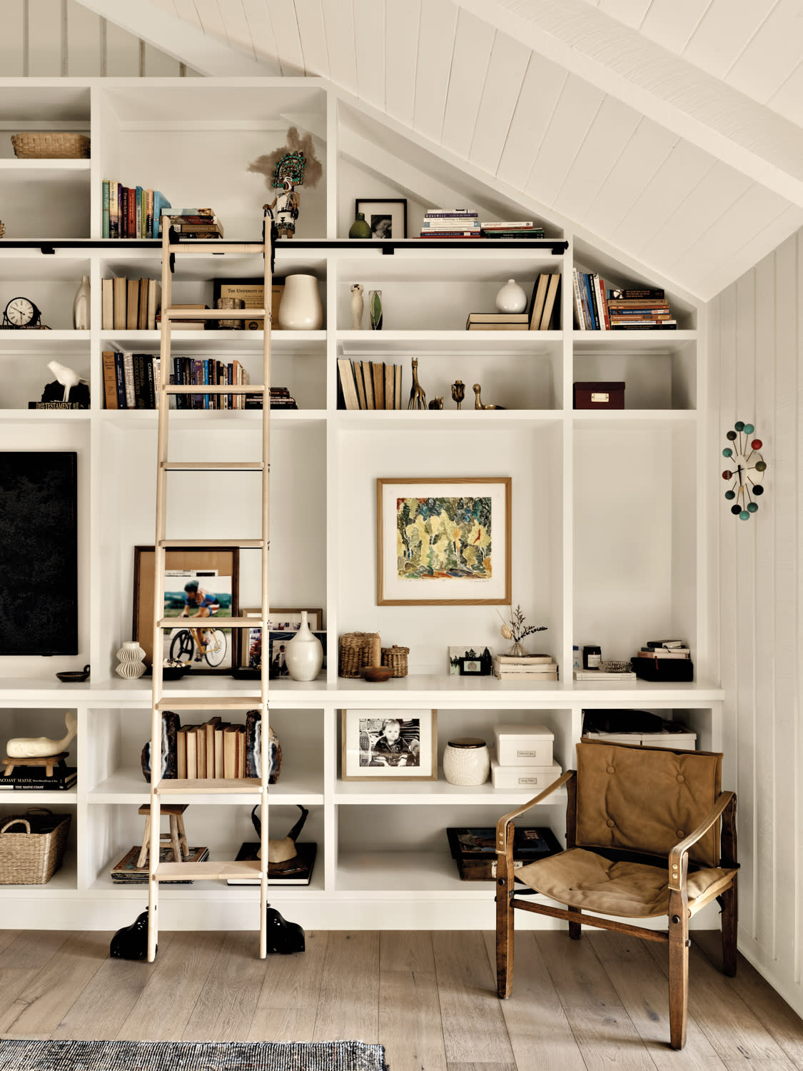 A ladder glides along floor-to ceiling built-in shelving in a white living room with shiplap walls