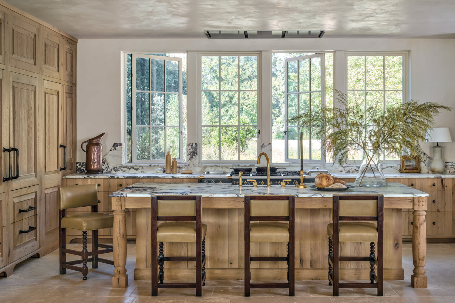 Spacious kitchen with light wood cabinets and large windows. An island with a marble countertop and bar stools faces the windows. Bright and airy.