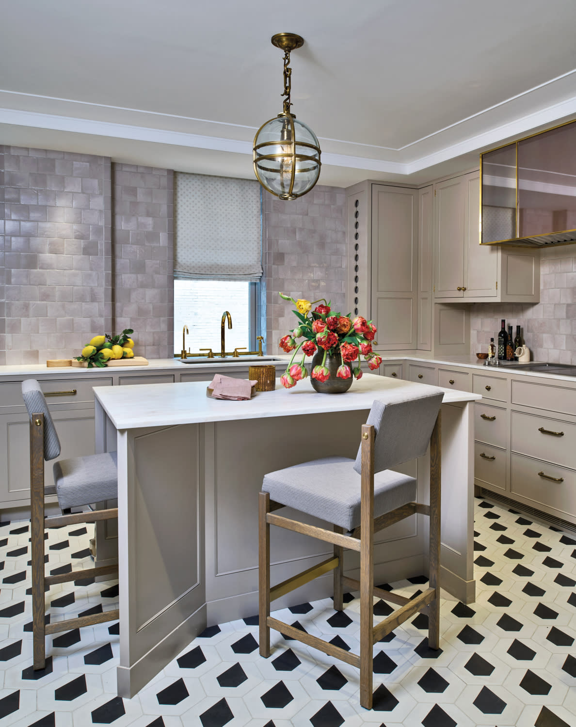 Elegant kitchen with gray cabinets, patterned black-and-white tile floor, and central island with two cushioned stools. A vase of tulips adds color.