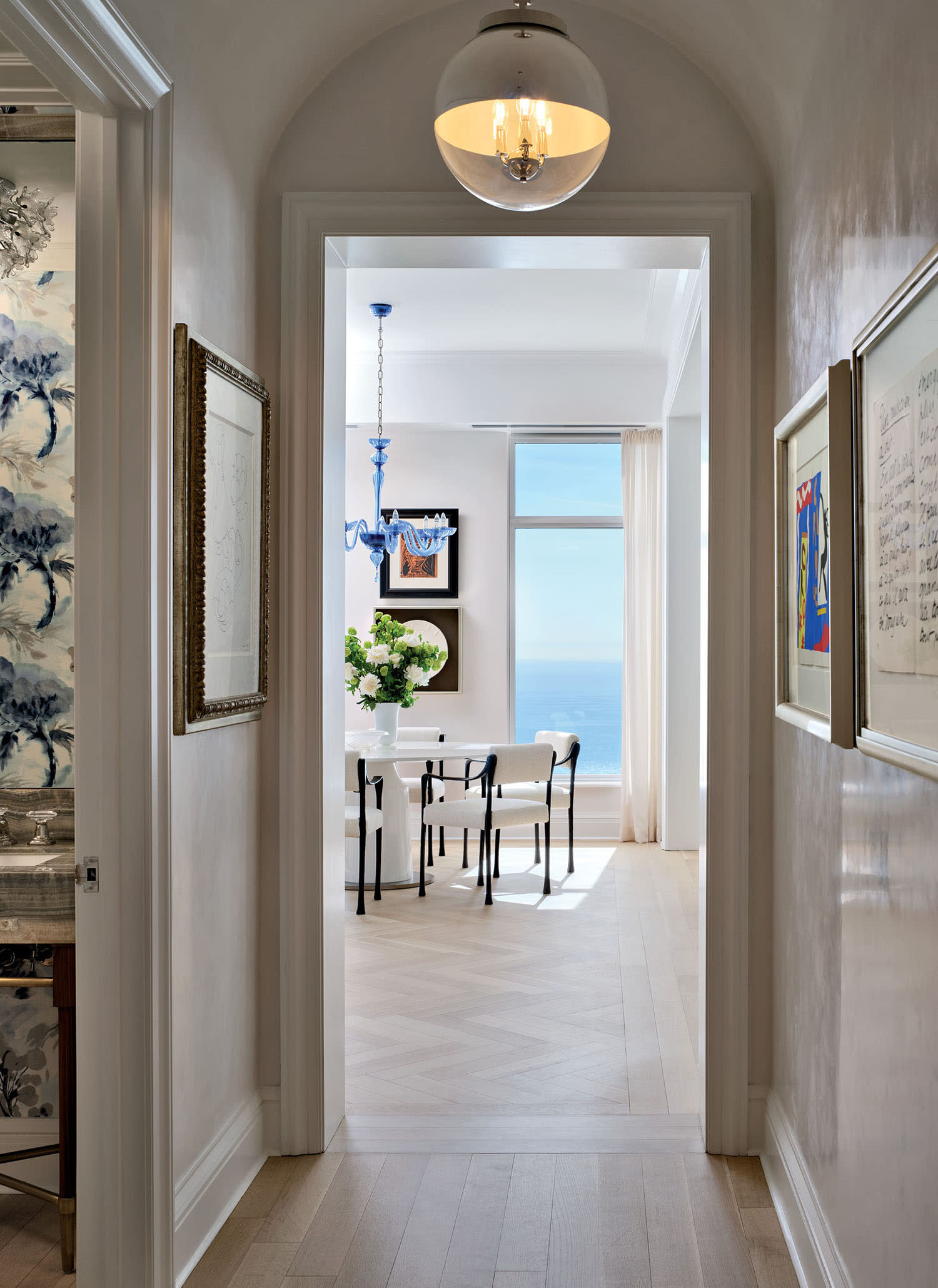 A hallway decorated with artwork and a round light fixture leading into a dining room space.