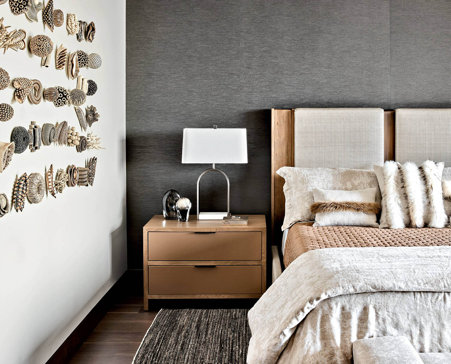 A bed with white and tan bedspread and accent pillows sits next to a nightstand near a wall sculpture.