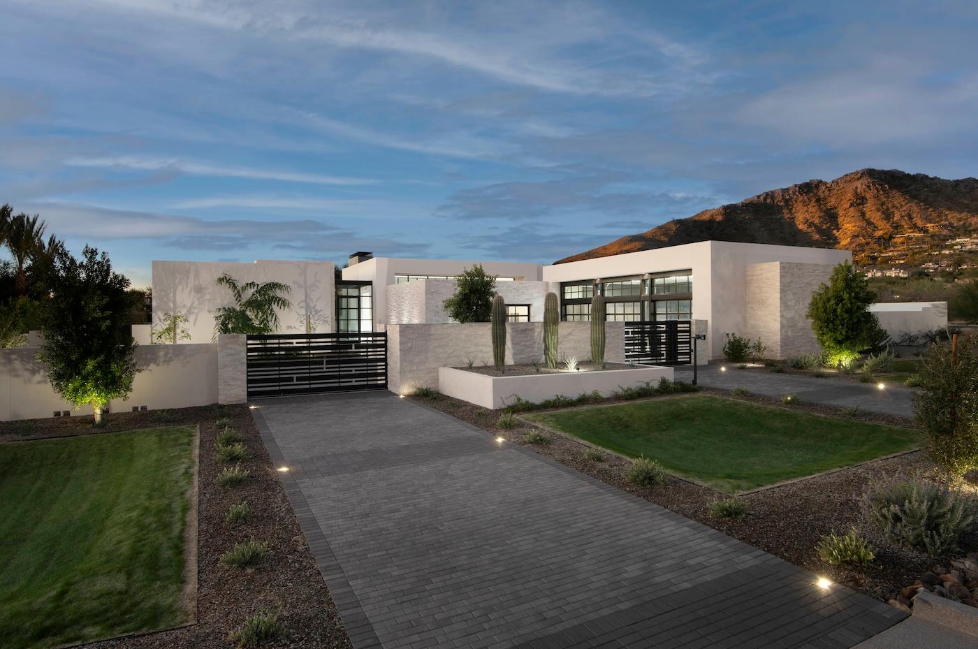Modern white house with large windows, minimalist landscaping, and a wide driveway. Surrounded by mountains under a blue sky with clouds at dusk.
