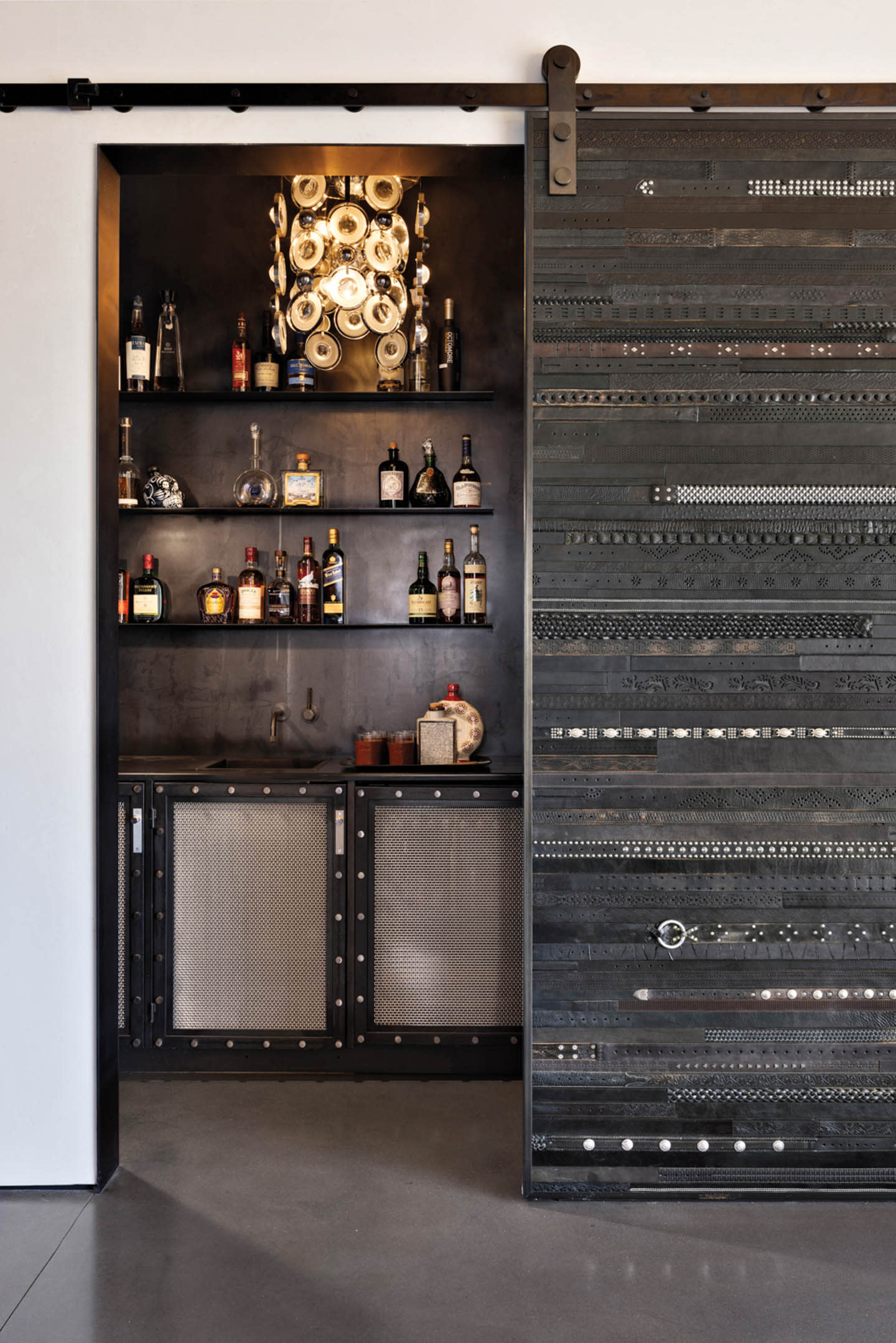 Industrial-style home bar with a black metal sliding door revealing shelves of assorted liquor bottles. Overhead lighting creates a cozy ambiance.