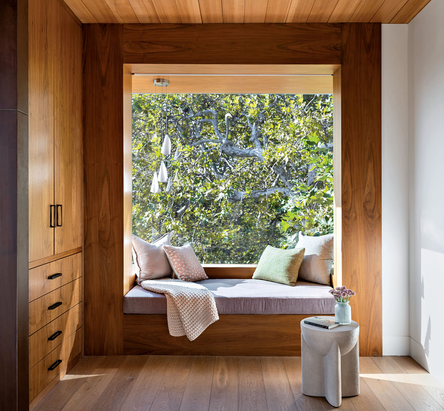 Cozy wooden alcove with a window seat adorned with pastel cushions and a throw, overlooking lush green trees