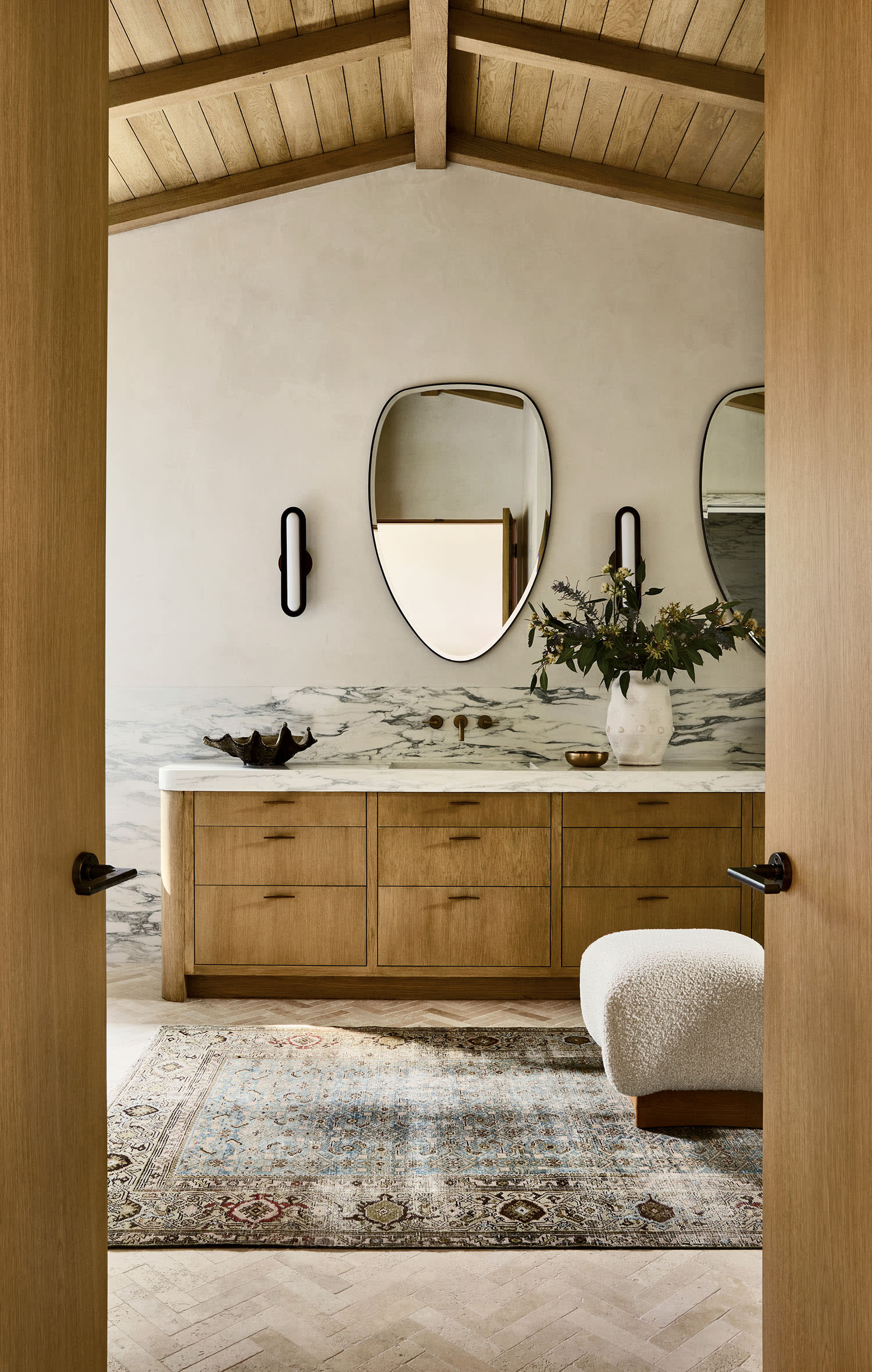 Spacious bathroom with wooden cabinets, marble countertop, and decorative mirrors. A plush stool and earthy-toned rug add warmth and elegance.
