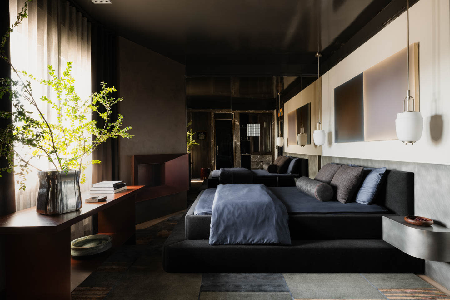 Sophisticated modern bedroom with dark gray walls, plush black sofas, pendant lights, abstract art, and a sunlit plant on a wooden console.
