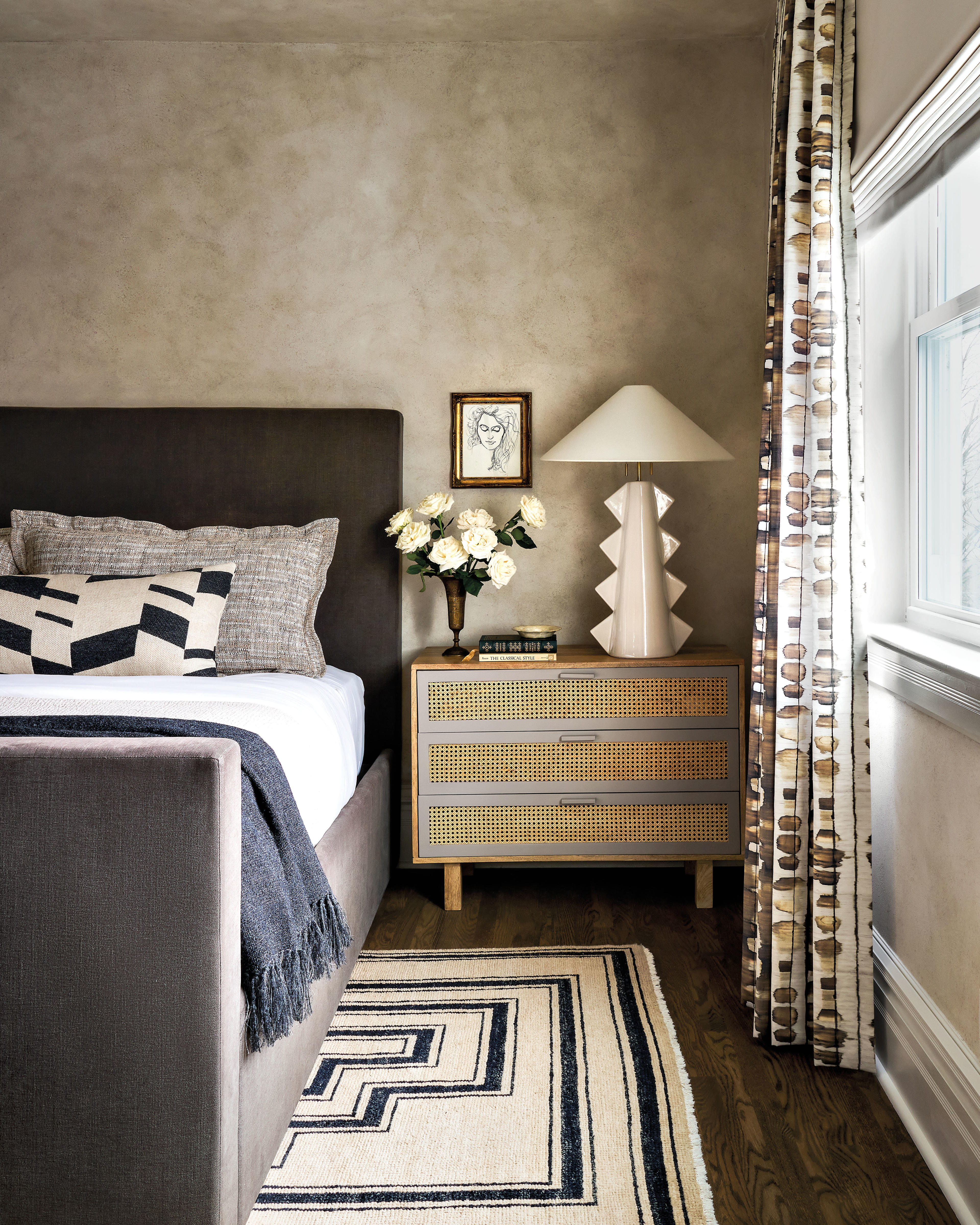 grey upholstered bed next to a cane side table in a beige limewashed bedroom