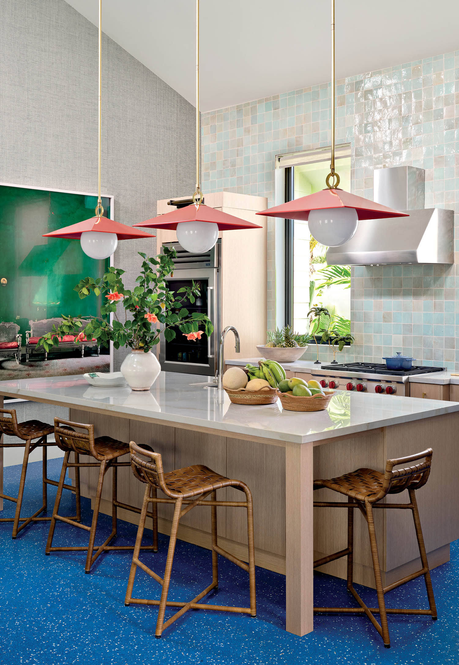 Modern kitchen with a blue speckled floor, light wood island, wicker stools, and red pendant lights. Green plants and fruit adorn the countertop, creating a fresh, vibrant atmosphere.