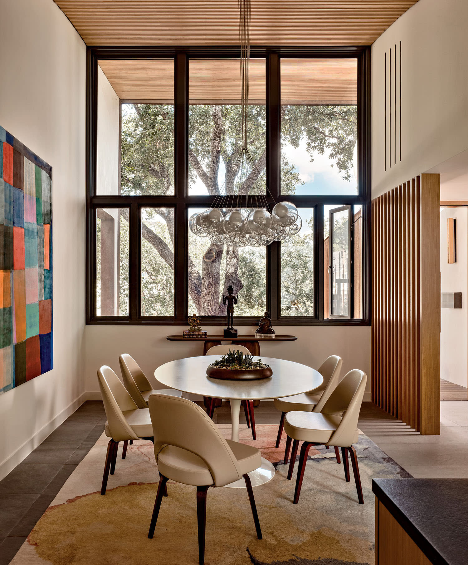 Modern dining room with a round table set for five, surrounded by beige chairs, beneath a bubble chandelier. Large window showcases a green tree.