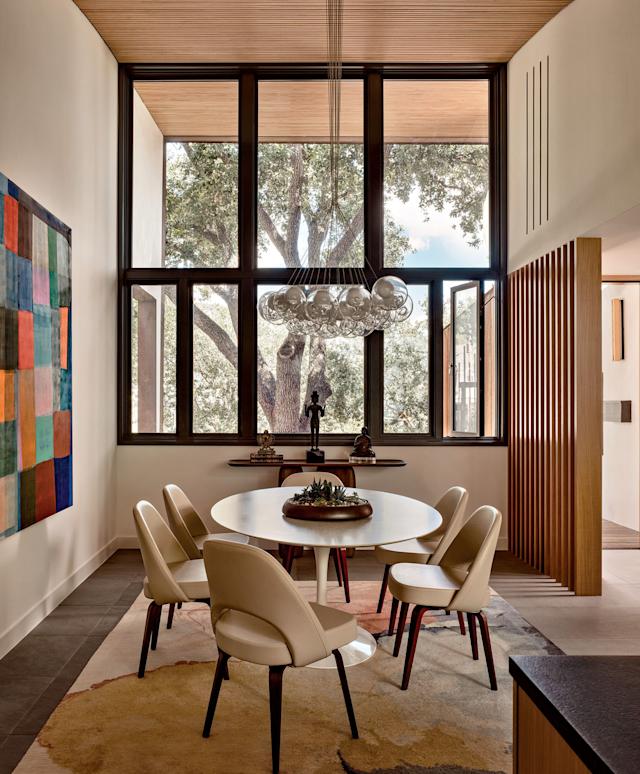 Modern dining room with a round table set for five, surrounded by beige chairs, beneath a bubble chandelier. Large window showcases a green tree.