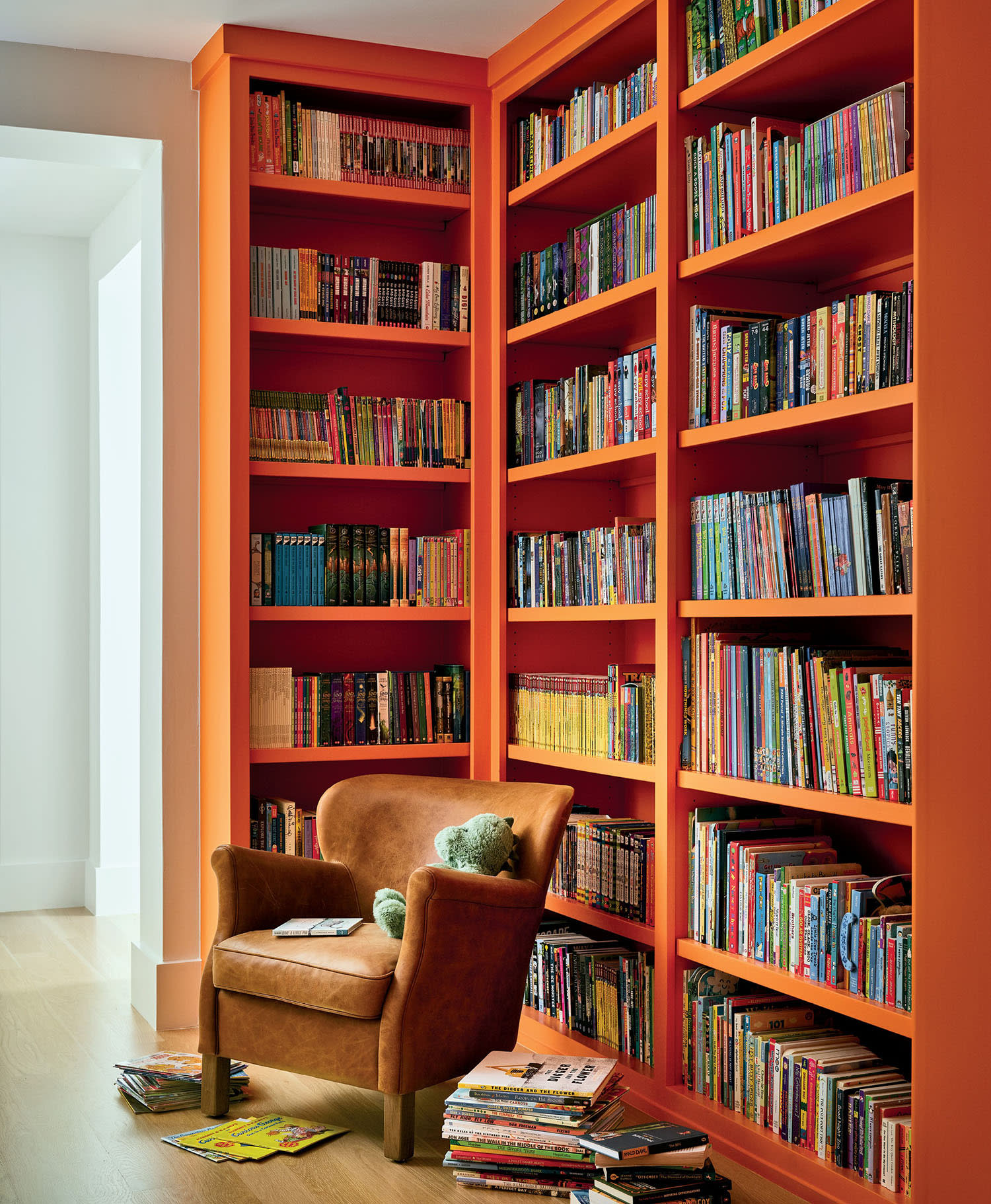 Cozy reading nook with an orange bookshelf filled with colorful books. A brown armchair holds a green teddy bear and a closed book. Stacks of books lie on the wooden floor nearby.