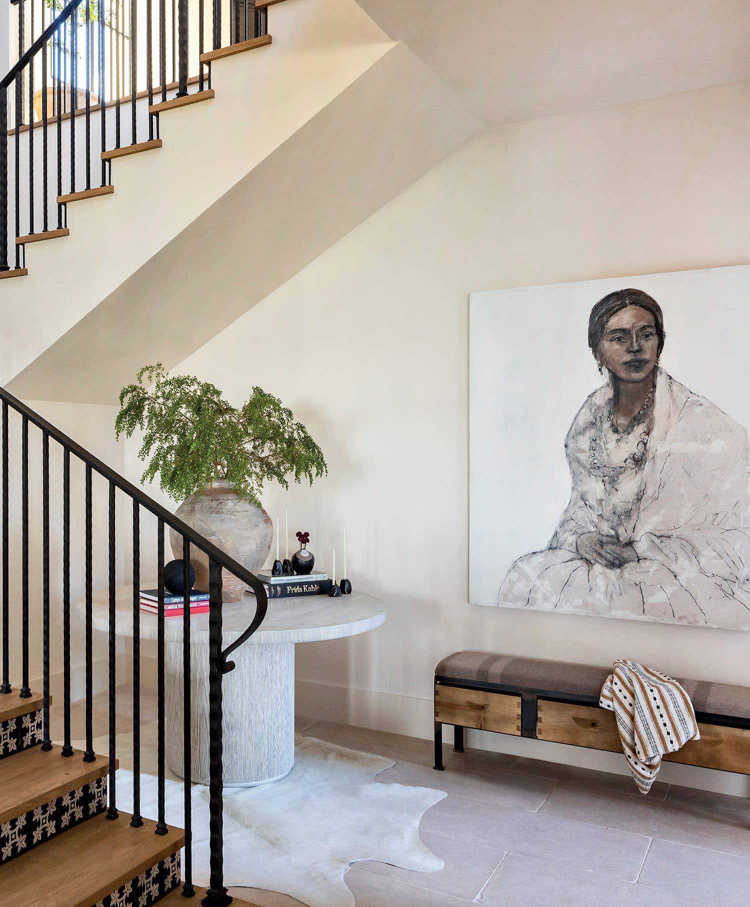 A stylish entryway with a modern staircase featuring black railings. A portrait of a woman, a round table with decor, and a bench with a draped blanket add warmth.