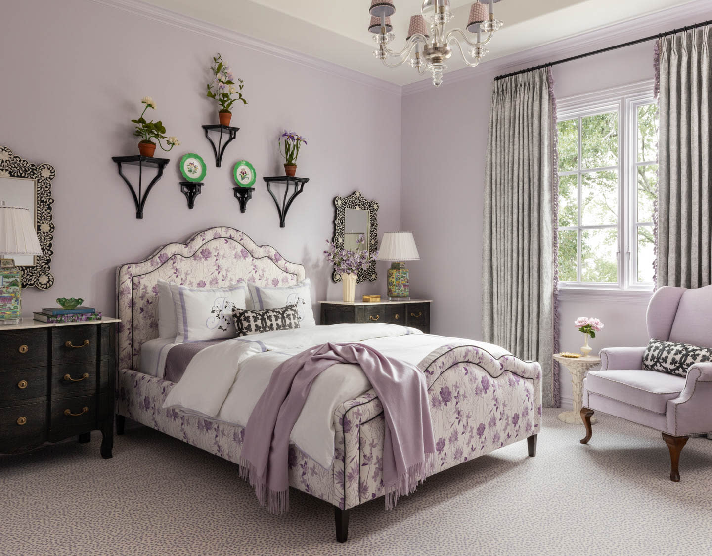 Elegant bedroom with lavender walls and a floral upholstered bed. Black nightstands, patterned mirrors, and wall plants enhance the cozy ambiance.