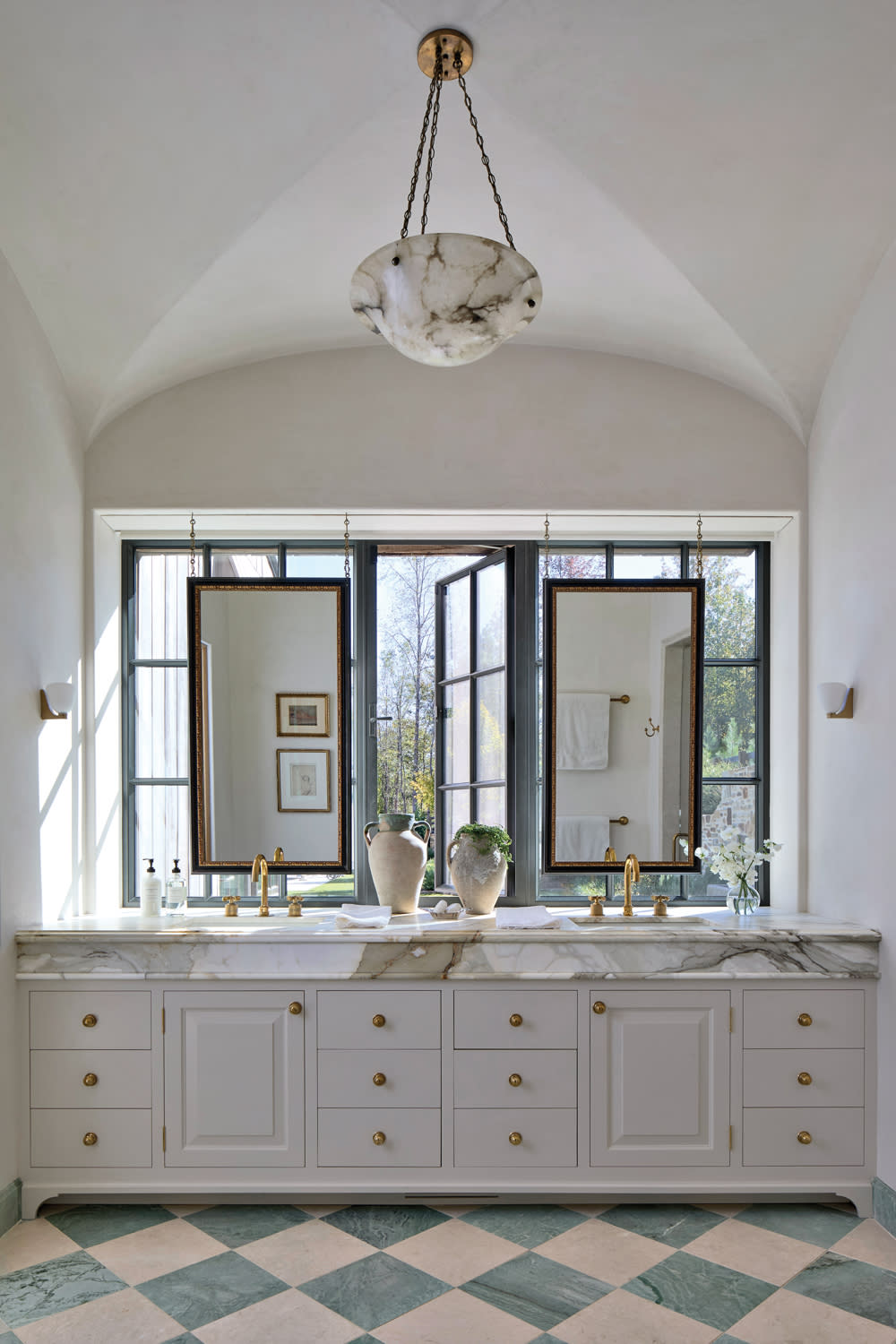 Elegant bathroom with a vaulted ceiling, a marble double sink, gold fixtures, two large mirrors, wide windows, and a checkerboard marble floor.