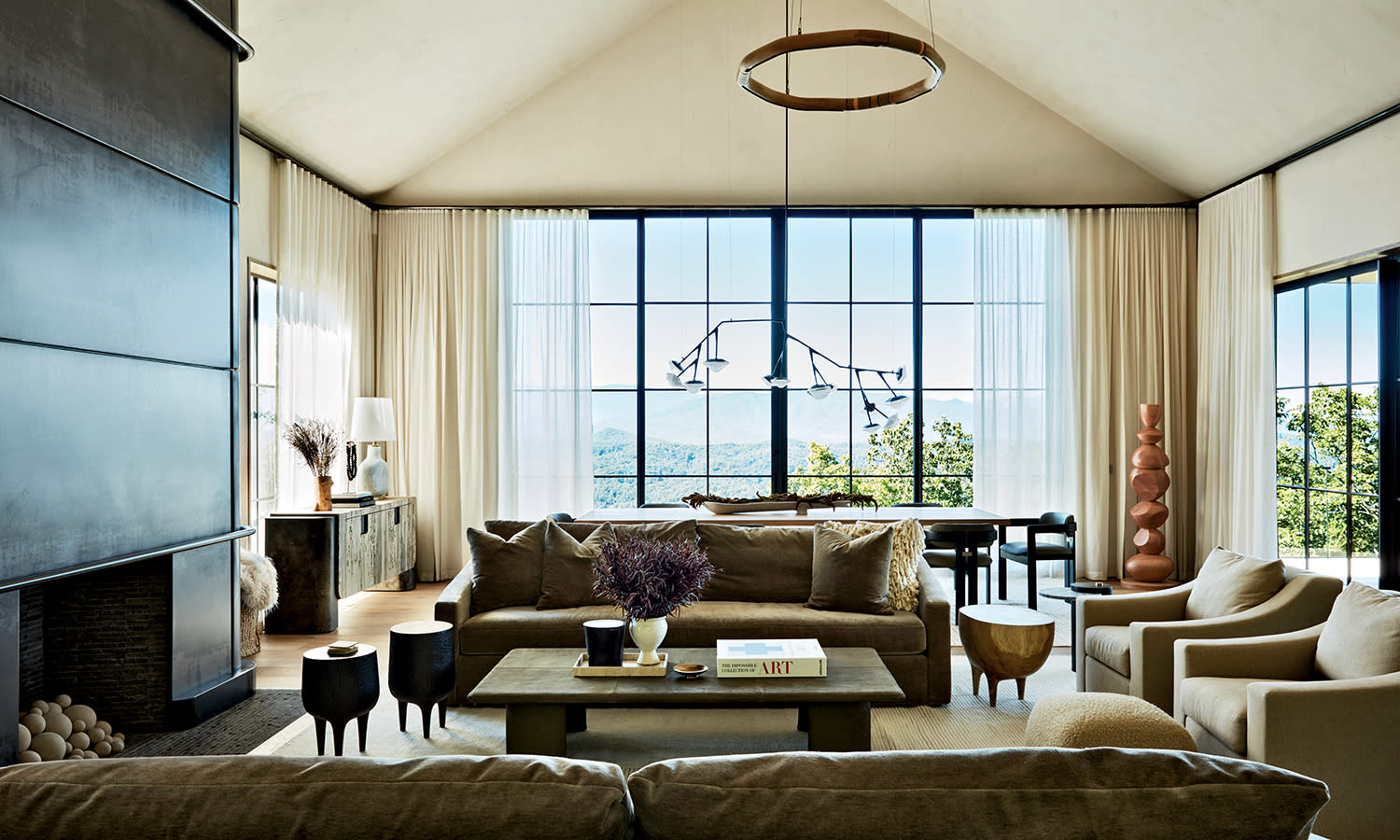 A dining room table sits nestled at the back of a living room, set with chairs and overlooking a large window with curtains.