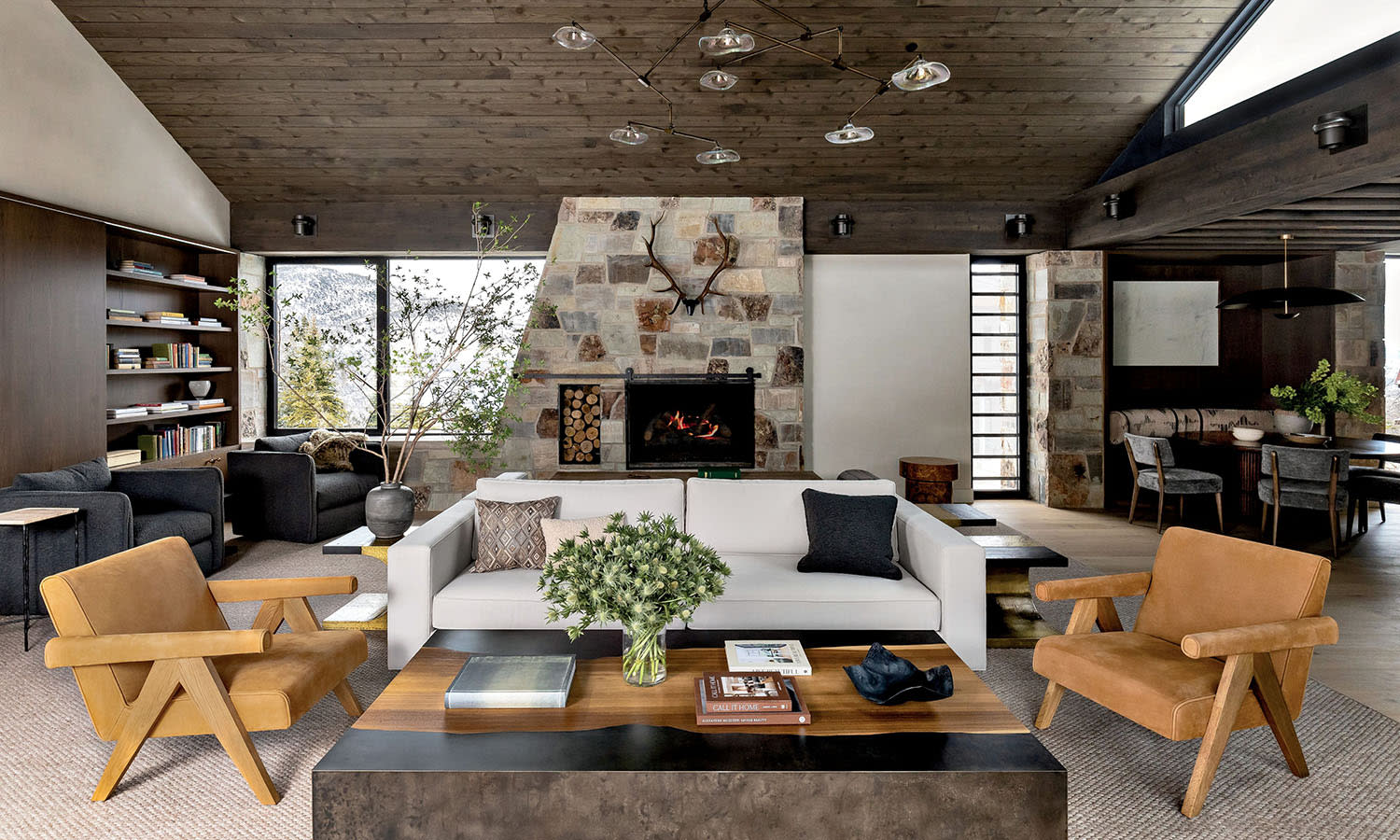 Sophisticated living room with stone fireplace, white sofa, brown armchairs, and wooden ceiling. Shelves, books, plants, and cozy ambiance.