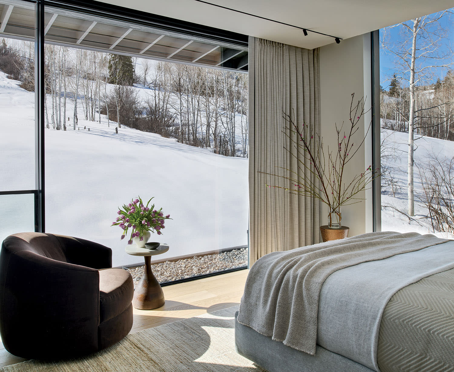 A serene bedroom with a large window view of a snowy landscape. The room features a cozy bed, a plush brown chair, a side table with flowers, and peaceful decor.
